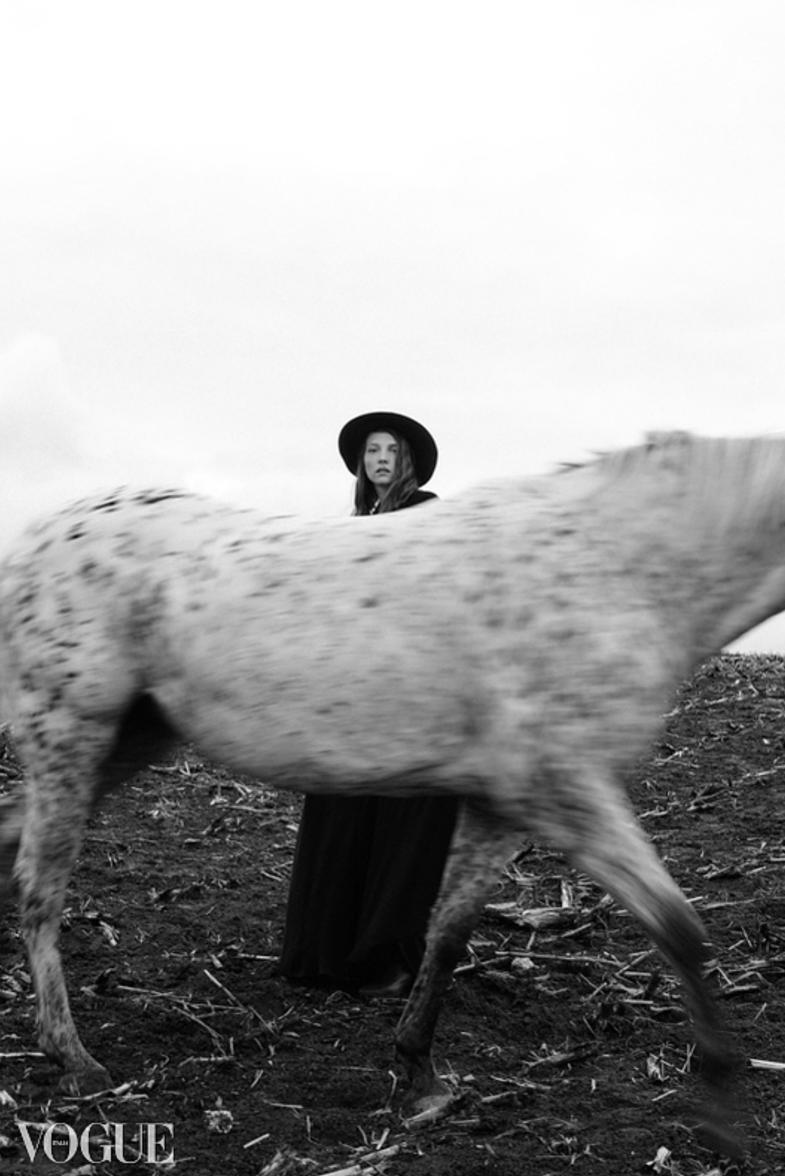 Black and white photo of a woman in a black dress and hat standing behind a spotted horse in a field.