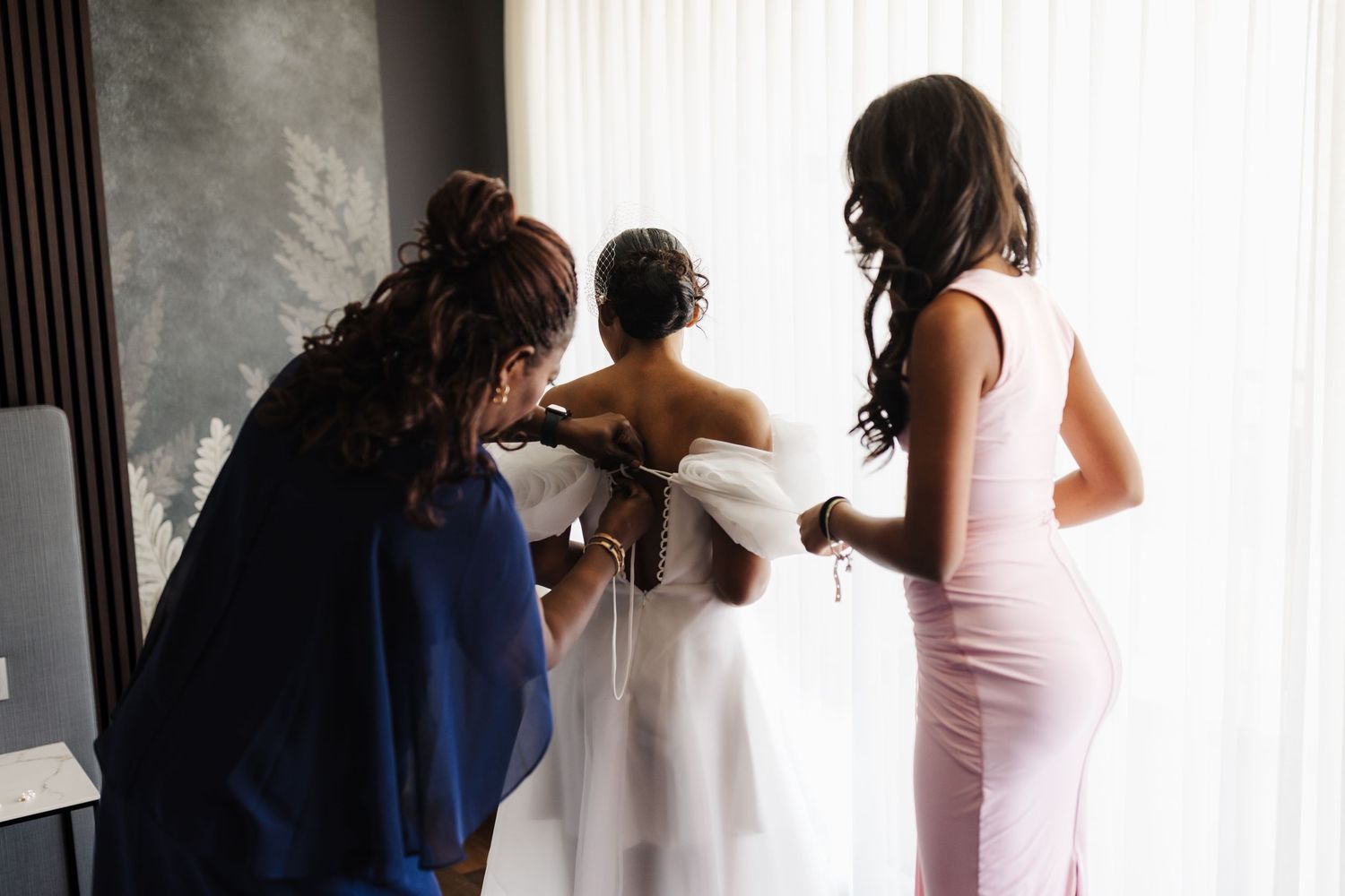 Two bridesmaids help a bride into her white off-shoulder wedding gown in a bright hotel room.