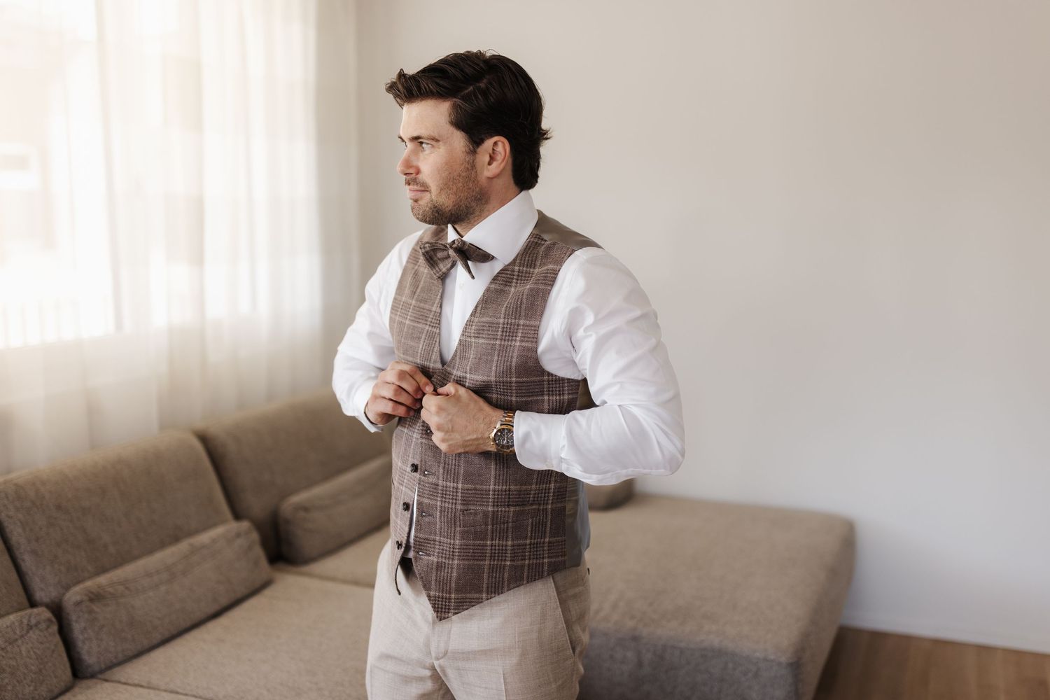 Elegant groom buttoning a brown plaid vest over a white shirt with a bow tie in a bright modern room.