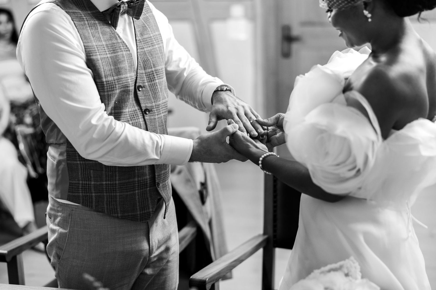 Black and white close-up of bride and groom holding hands during wedding ring exchange ceremony.
