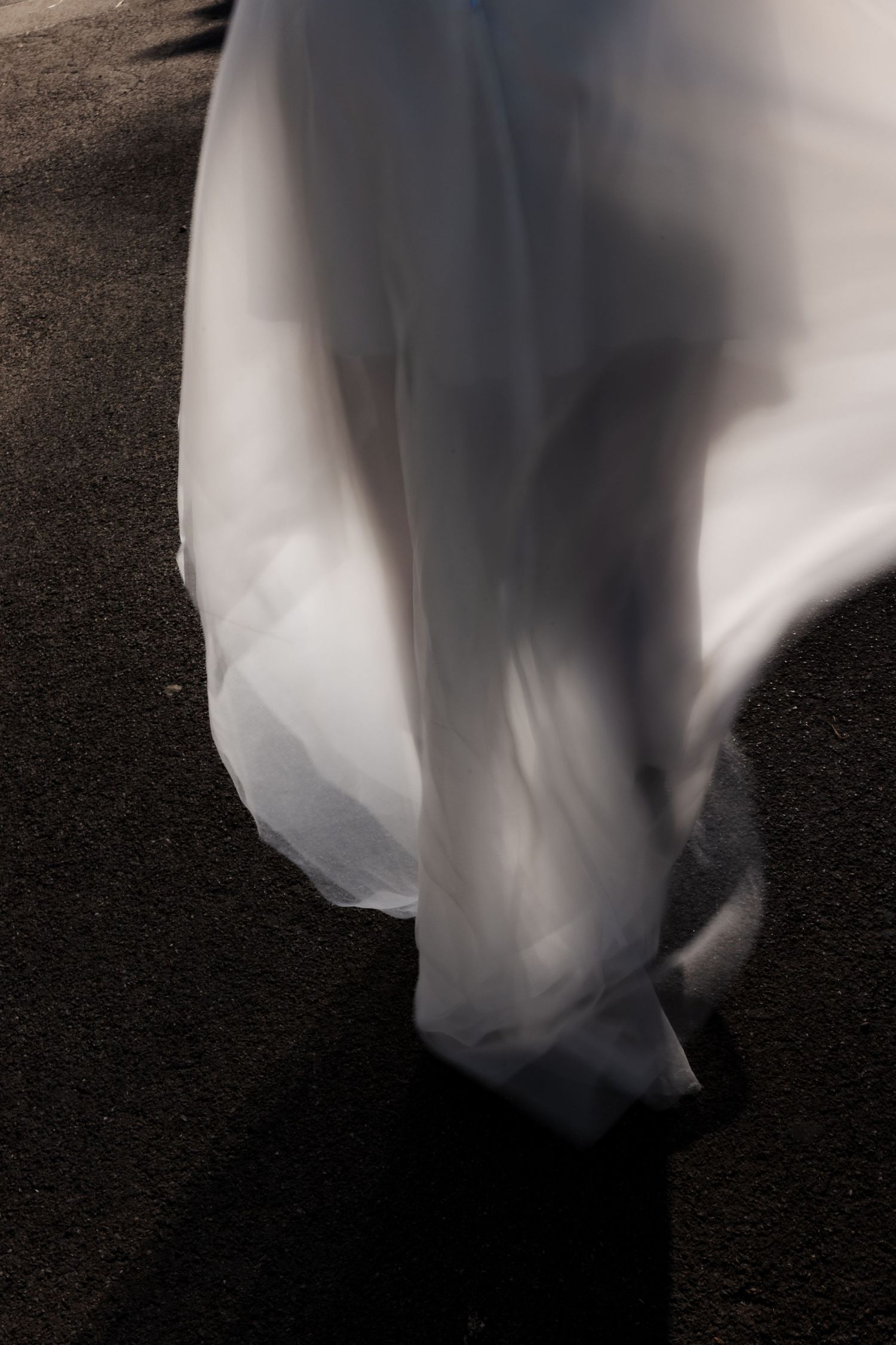 Artistic motion blur of white wedding dress train sweeping across dark pavement.