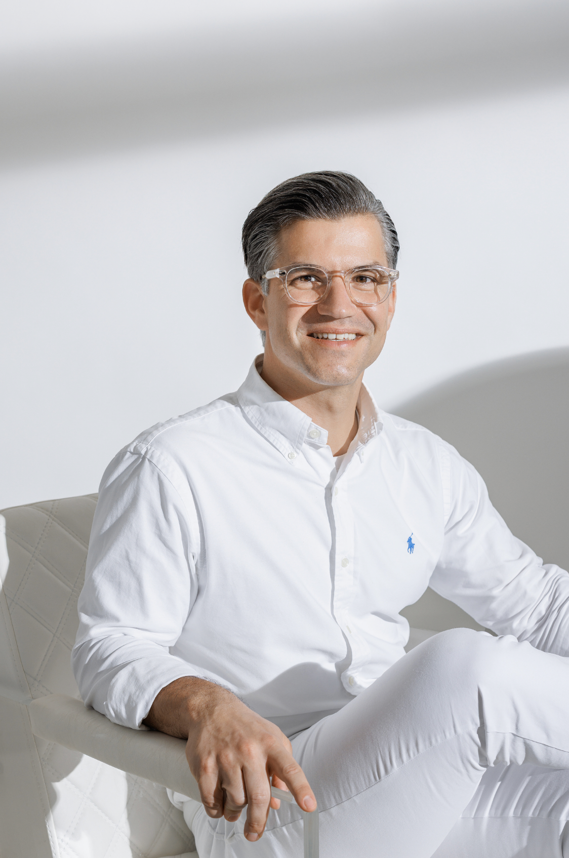 A smiling man wearing glasses and a white Ralph Lauren shirt sits relaxed in a modern chair.