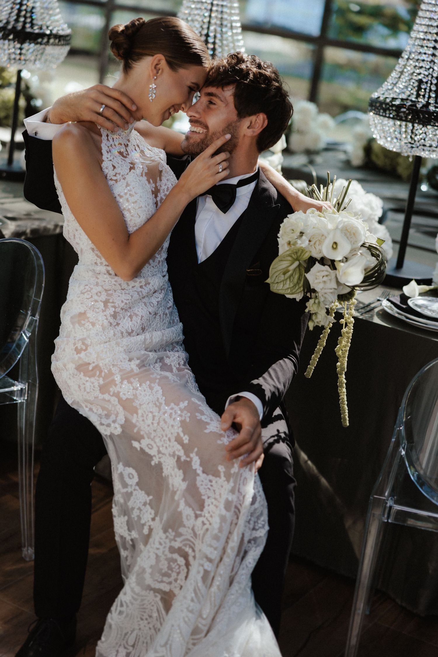 Bride in lace gown sits on groom's lap, both smiling intimately at elegant wedding reception.