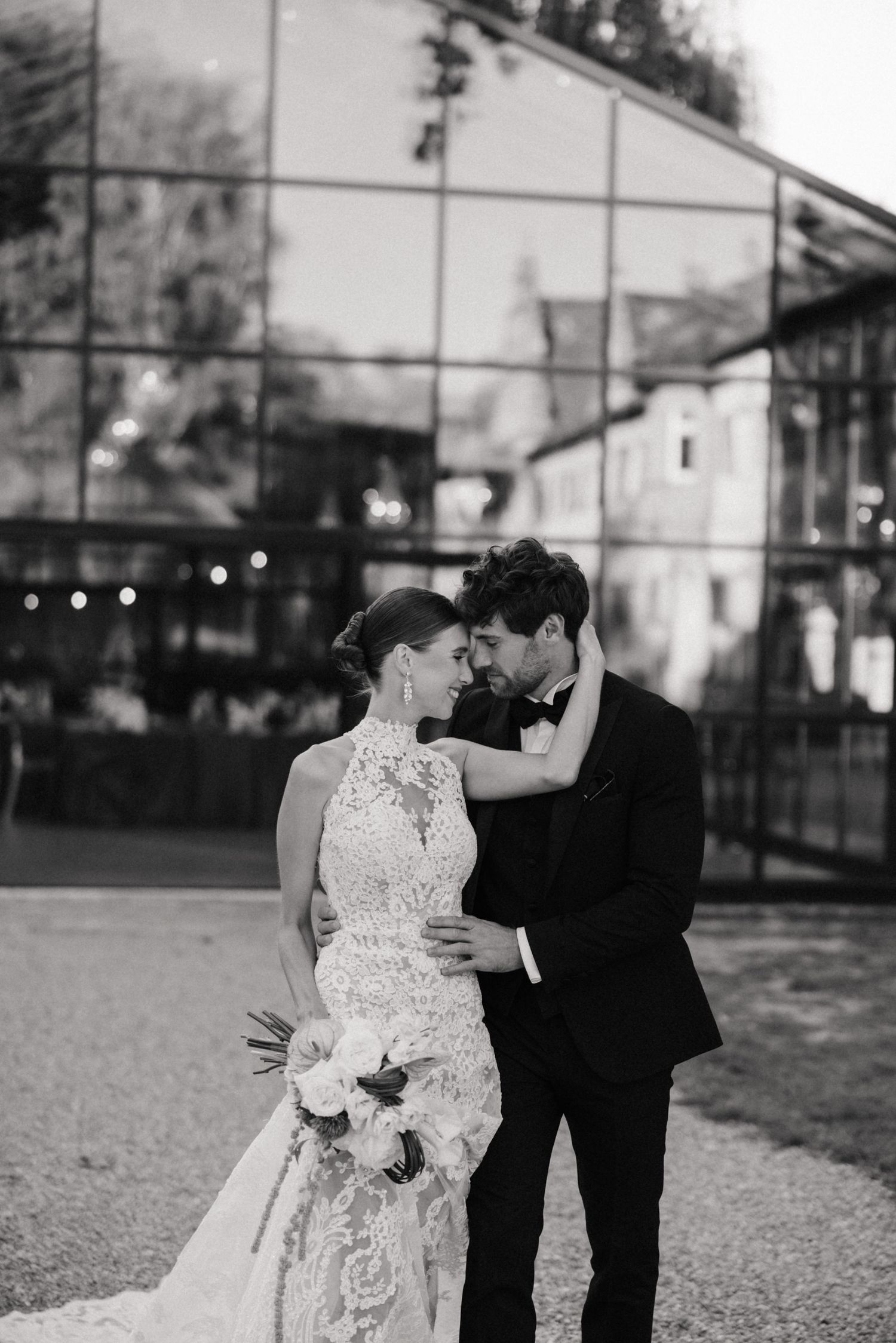 Black and white photo of bride in lace halter gown and groom in tuxedo embracing before glass building.
