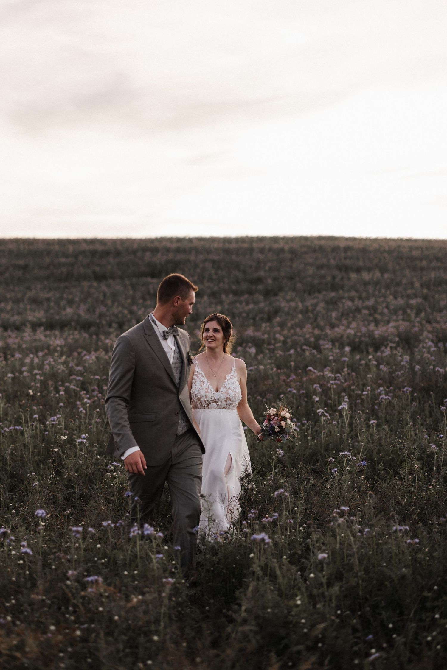 Smiling bride in lace gown and groom in gray suit walk through a wildflower field at dusk.