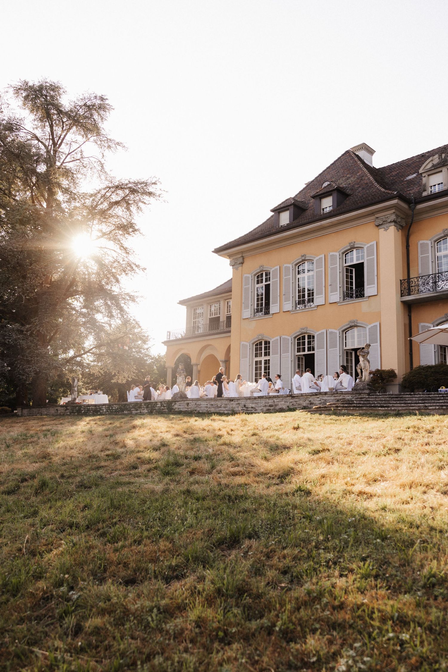 Golden sunset illuminates a yellow chateau with wedding guests gathered on the terrace steps in white attire.