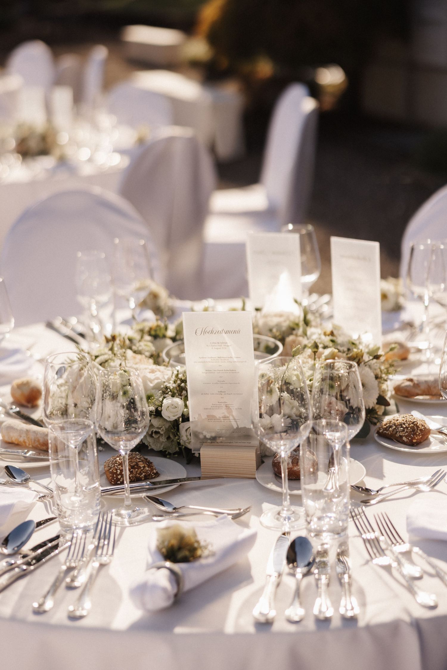 An elegant white wedding table setting features a floral wreath centerpiece, crystal glasses, and a printed menu card.