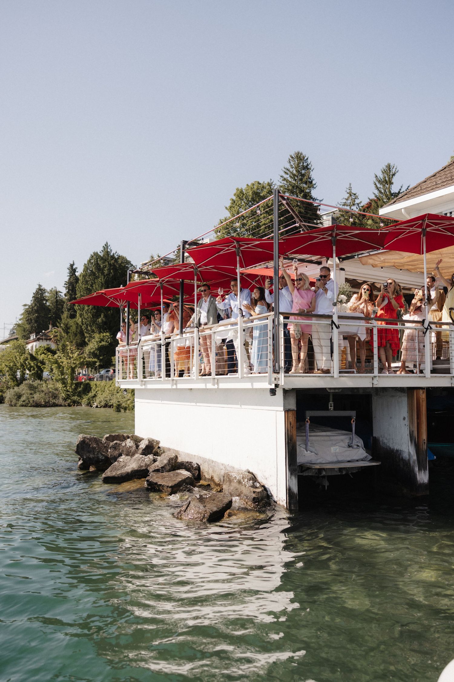 Wedding guests gathered on a lakeside restaurant deck with red umbrellas overlooking calm blue water.
