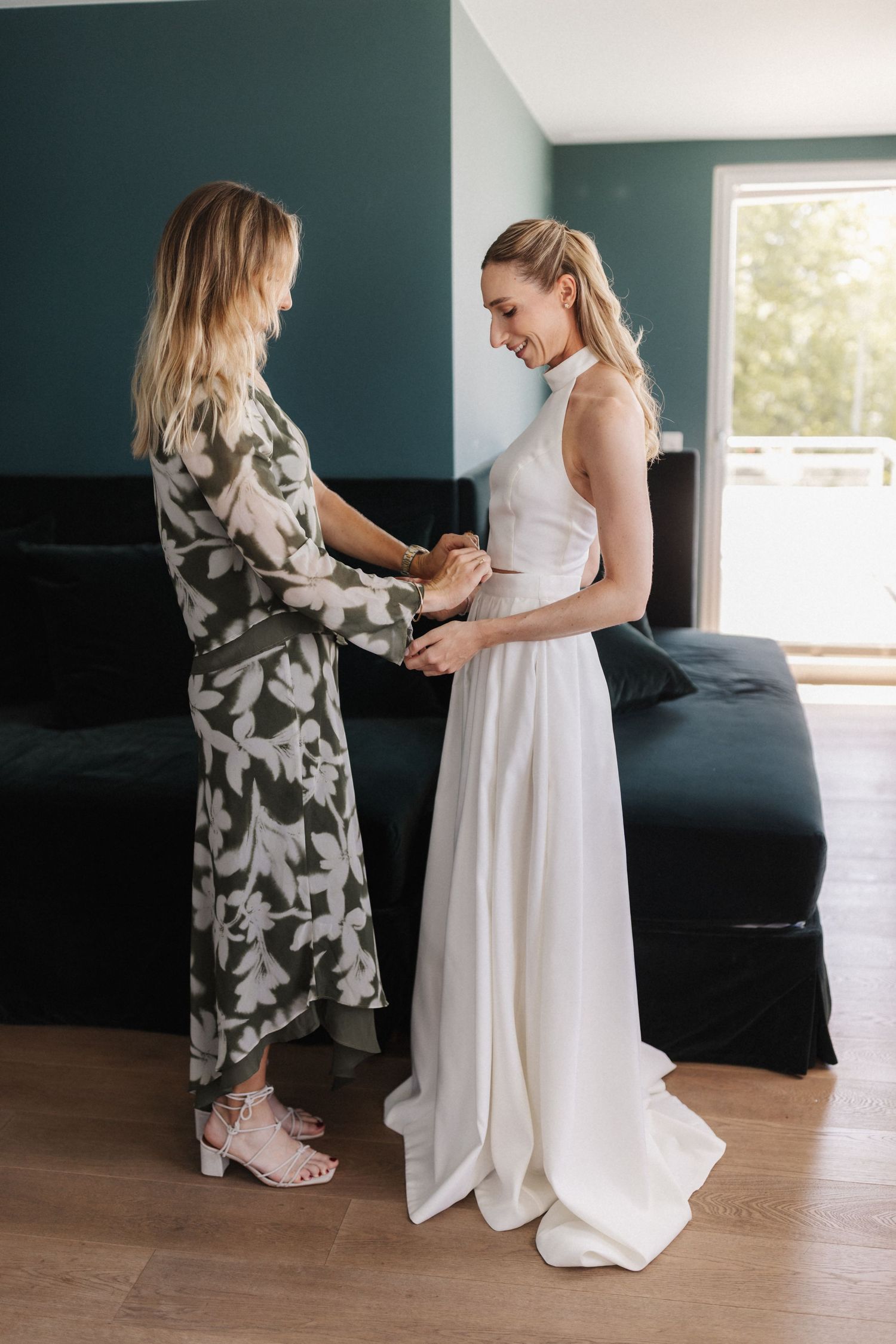 A woman in a floral dress helps a bride in a white halter gown with her outfit in a modern room.
