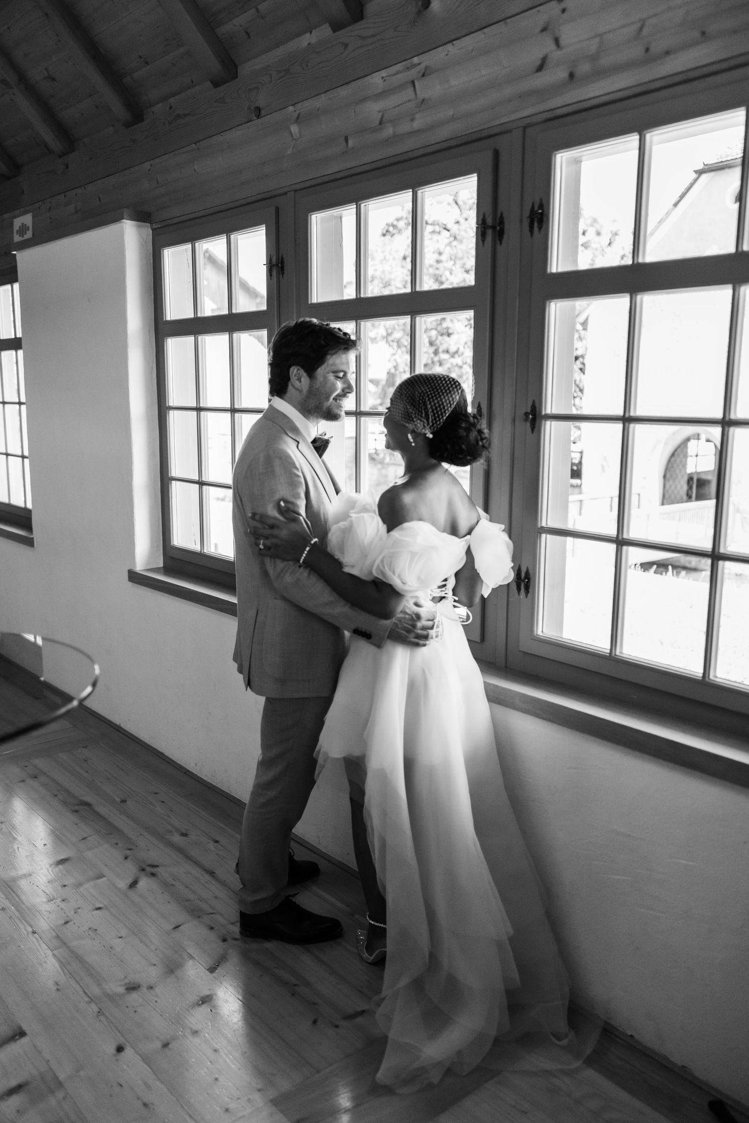 Black and white photo of groom embracing bride from behind near large windows indoors.