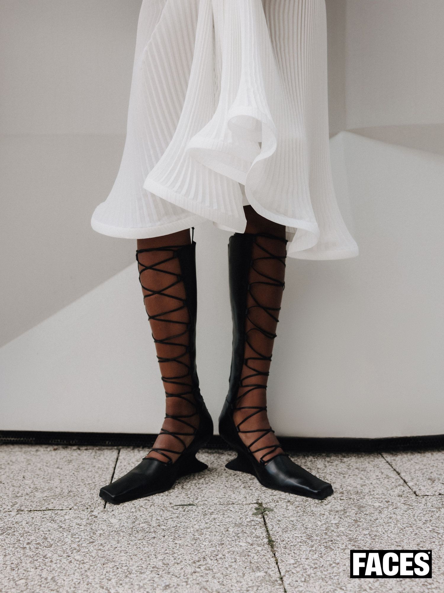 Close-up of black lace-up knee-high boots worn with sheer tights under a white pleated skirt hem.