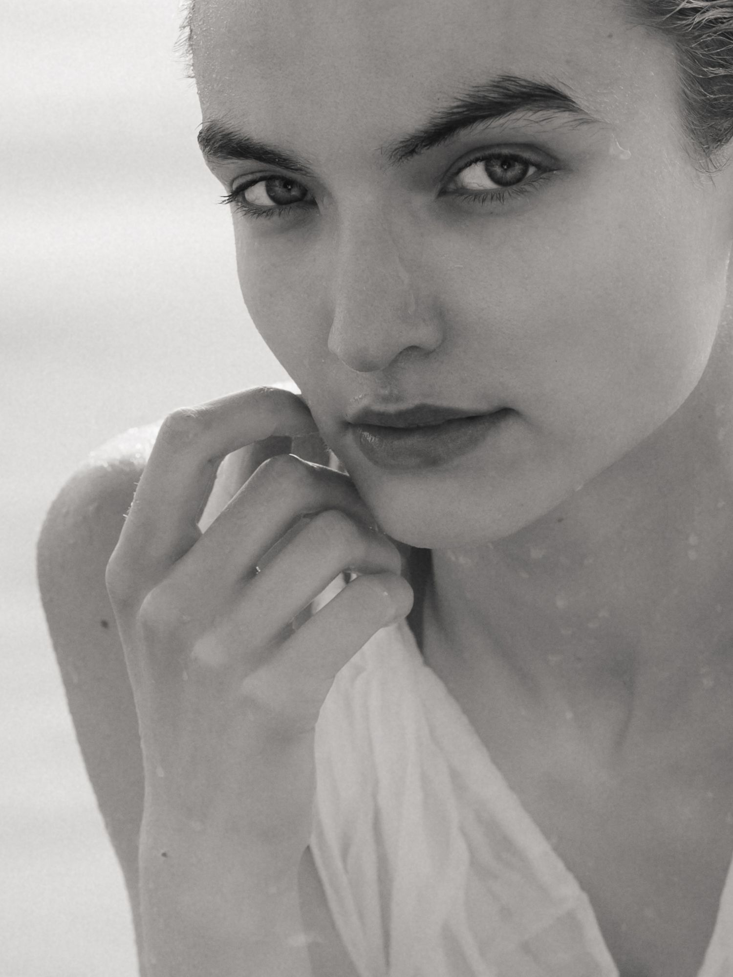 Black and white close-up portrait of woman with striking eyes holding hands near face wearing white top.