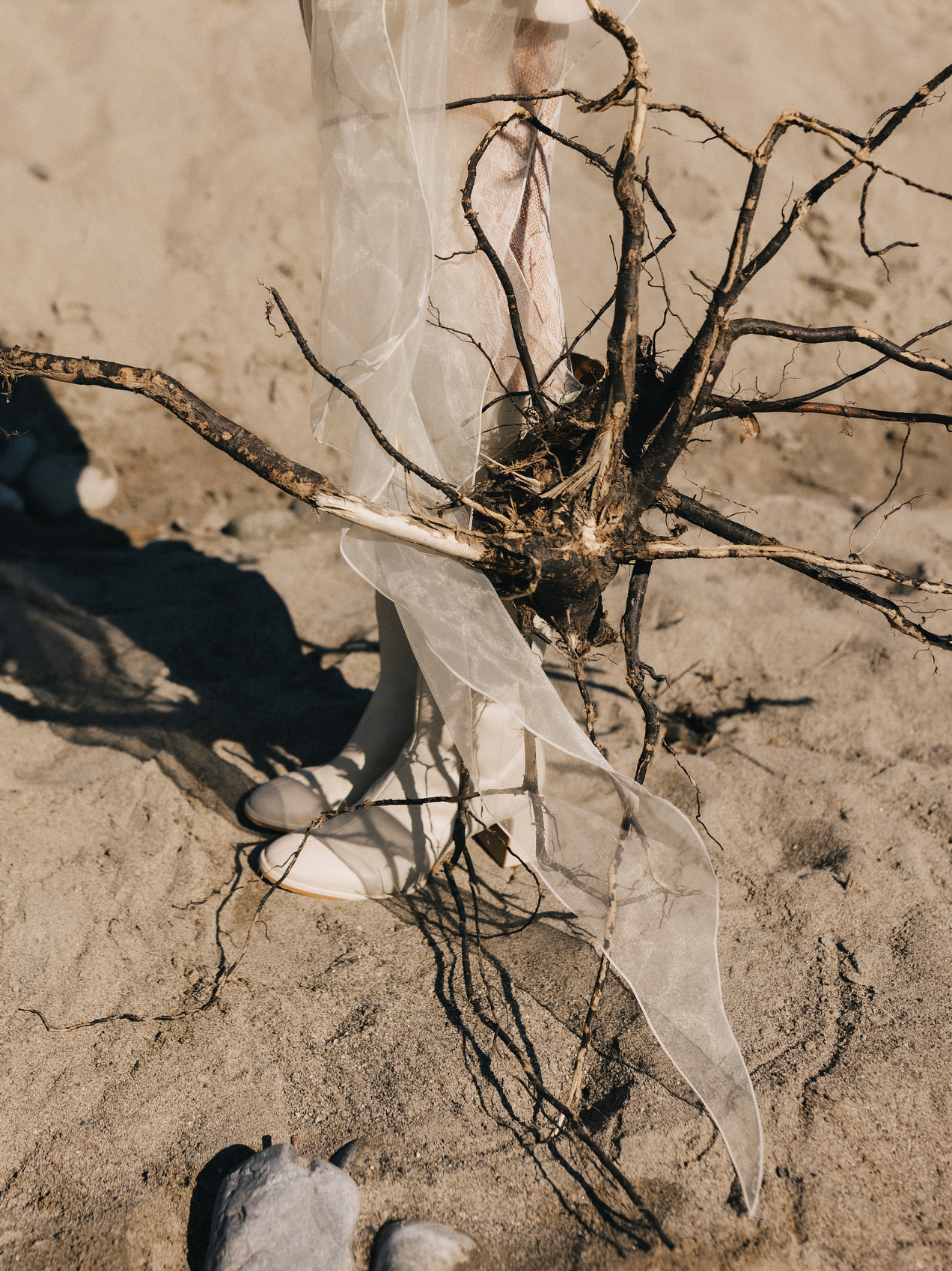 Delicate white fabric draped over dry twisted branches on sandy beach ground.