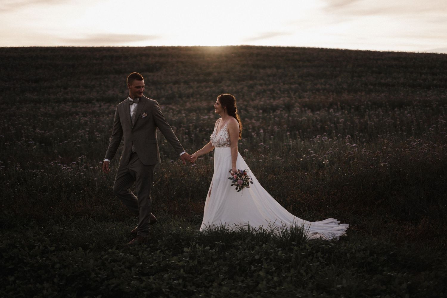 Bride and groom walk hand in hand through a lush green field during a breathtaking golden sunset.