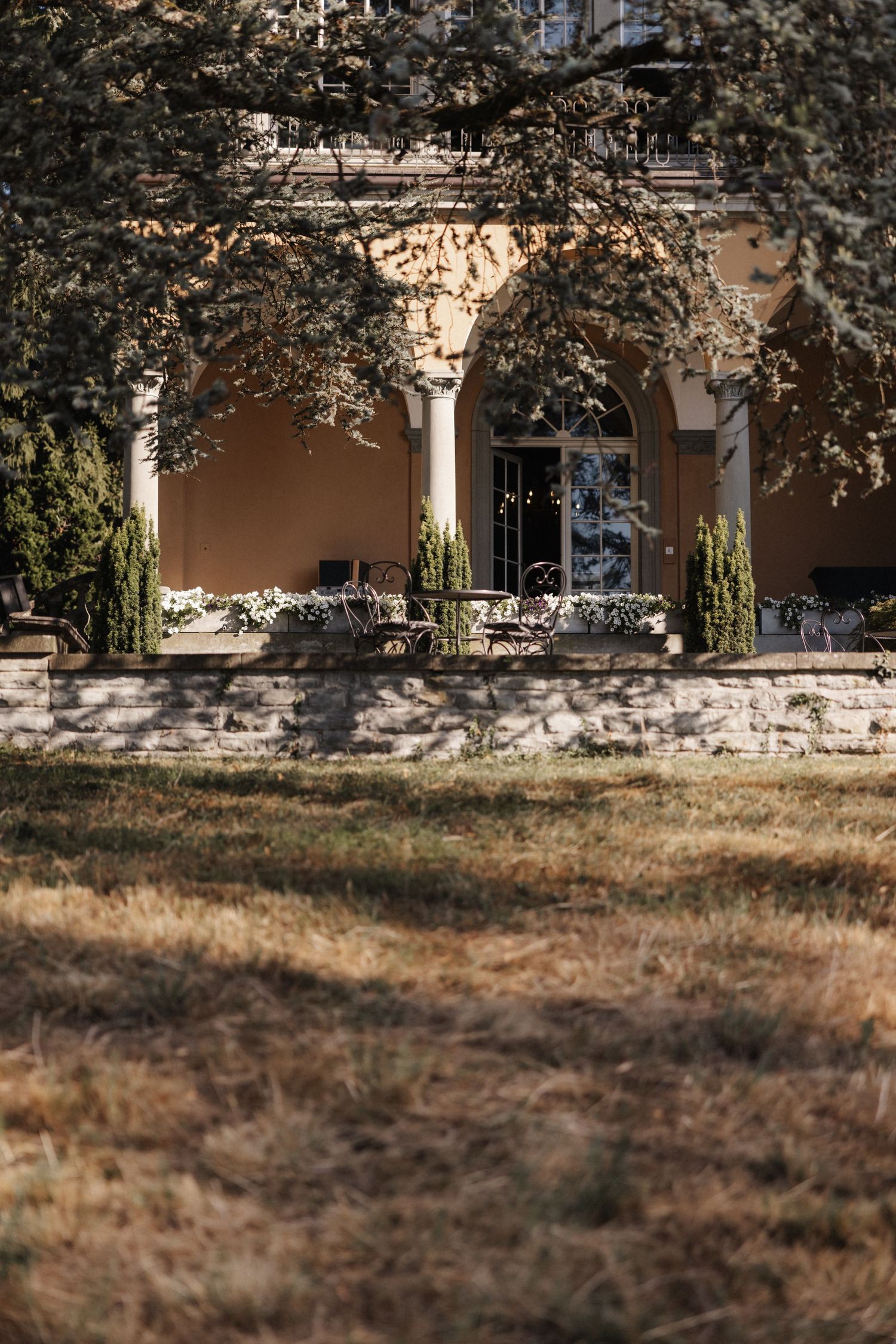 Rustic terrace of a beige stone manor with arched columns viewed through autumn tree branches.