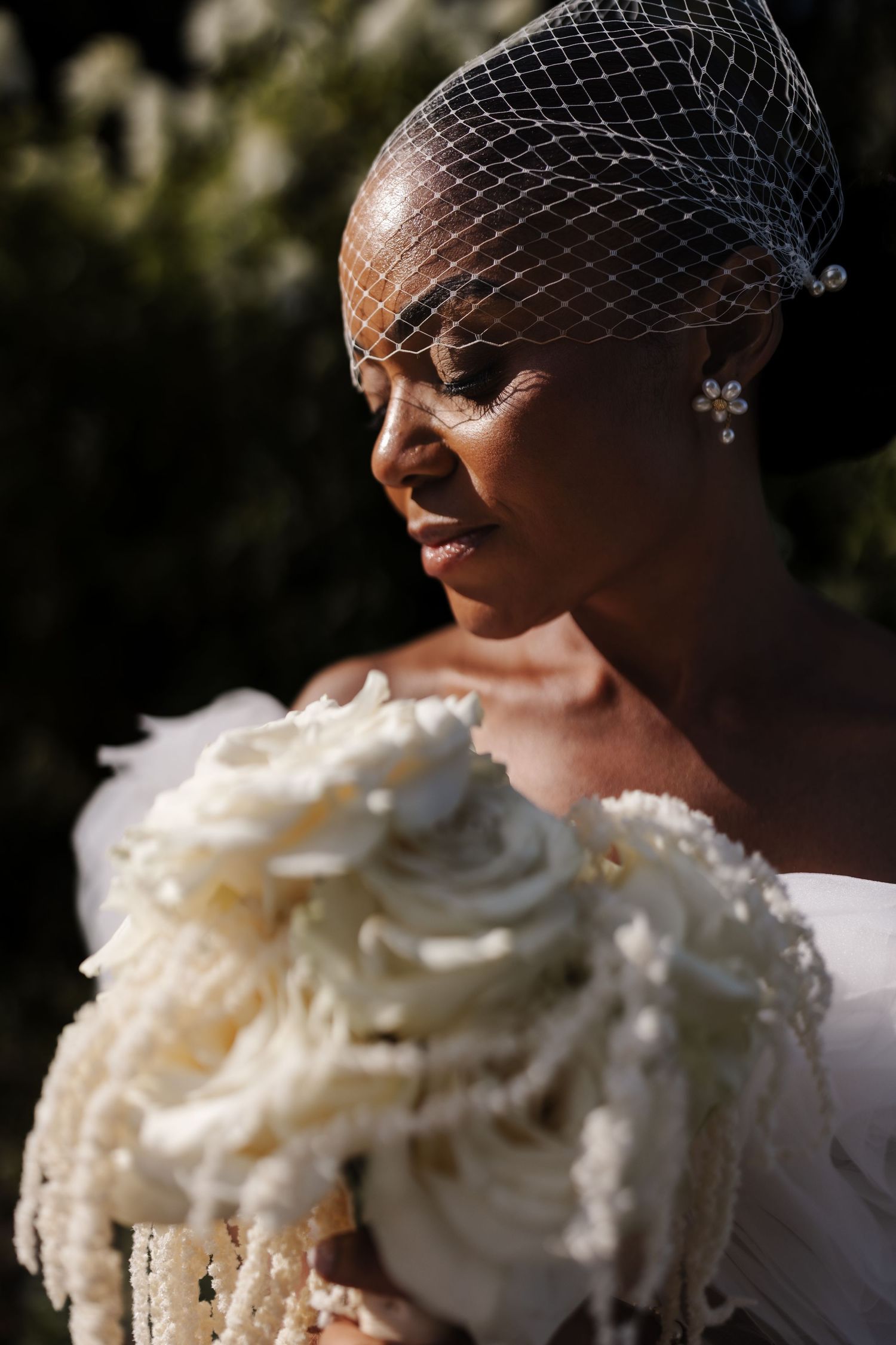 A bride in a birdcage veil gazes down at her white rose and pearl bridal bouquet in natural sunlight.