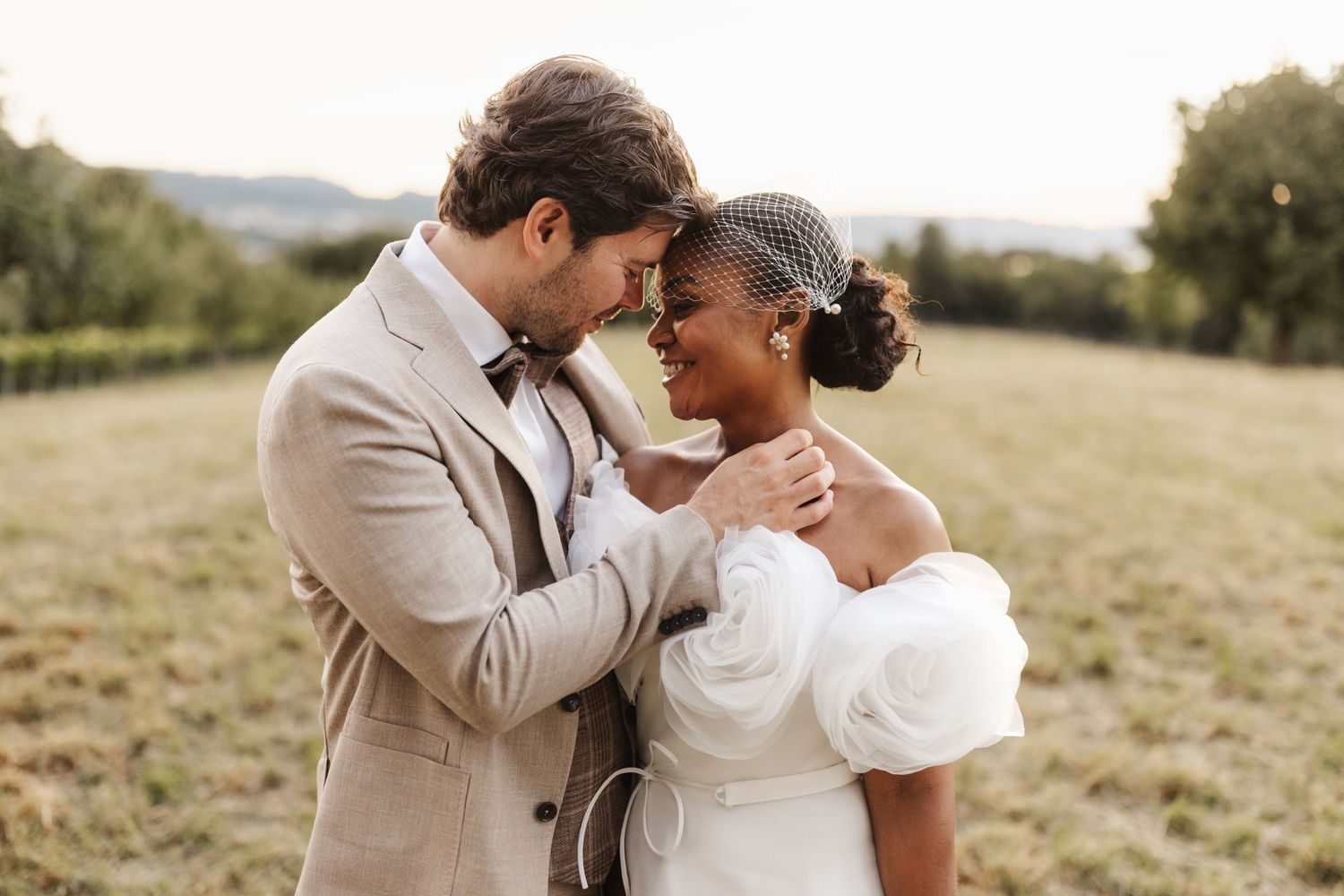 Interracial bride and groom sharing tender moment in sunlit field at golden hour.