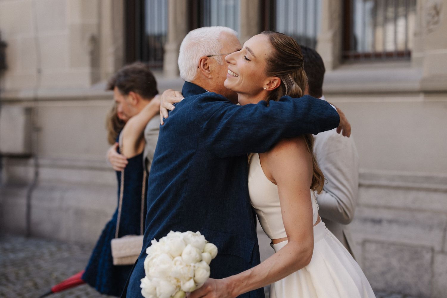 Bride in white gown embraces older gentleman outside European building while holding white peony bouquet on wedding day.