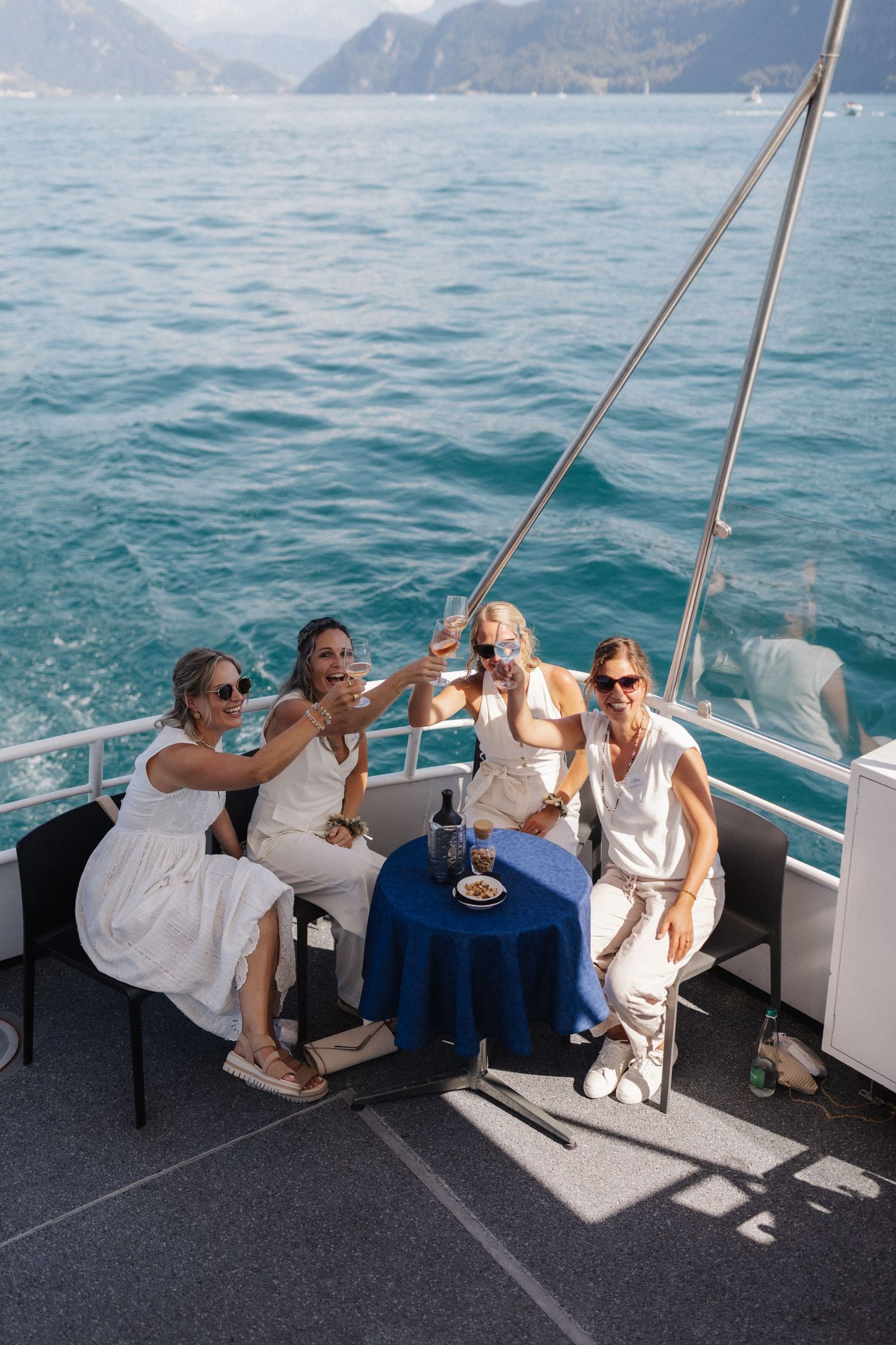 Four women in white dresses toast champagne glasses on a yacht deck with turquoise lake and mountains behind them.