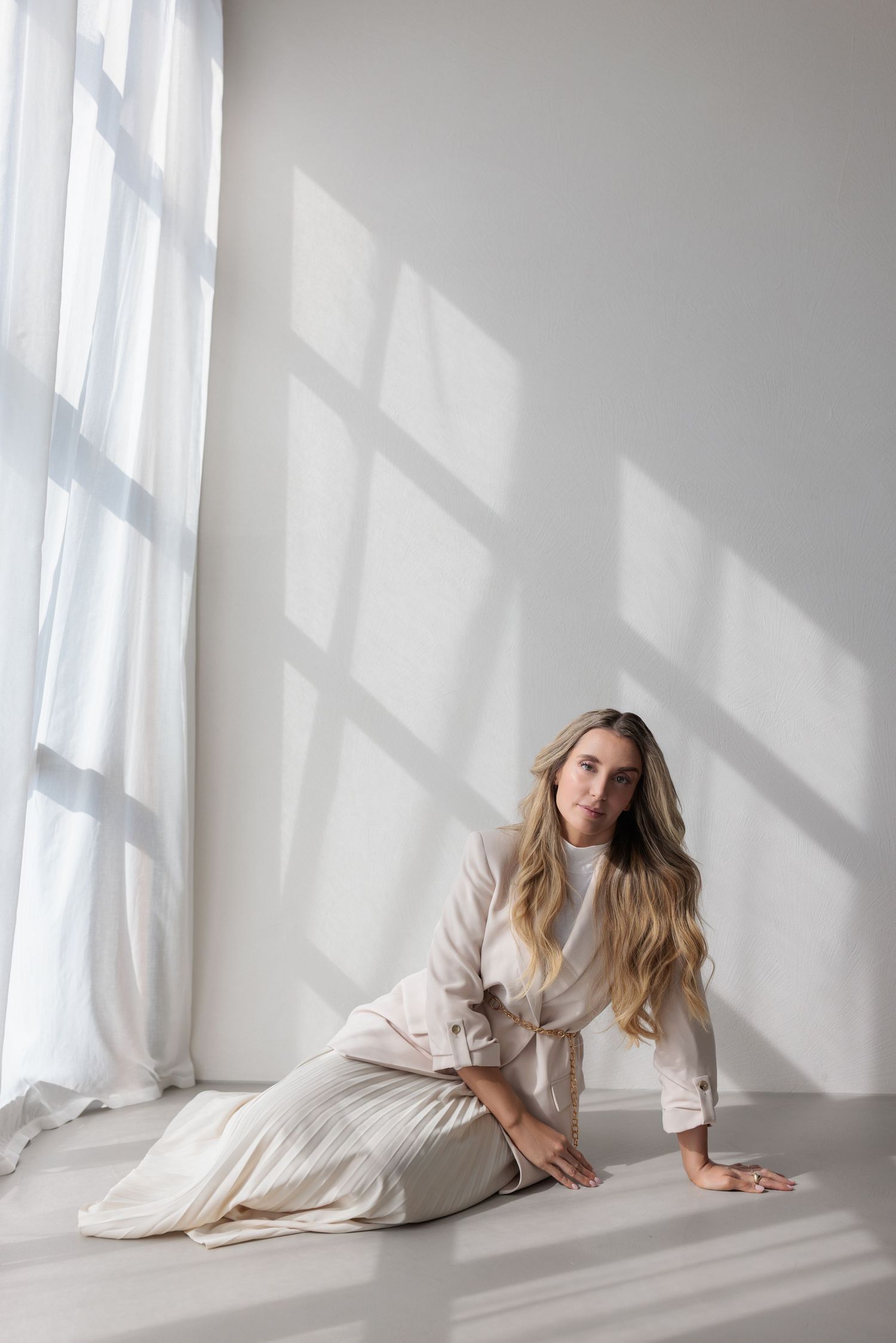 Blonde woman in beige outfit sitting gracefully on white floor near curtained window with natural light.