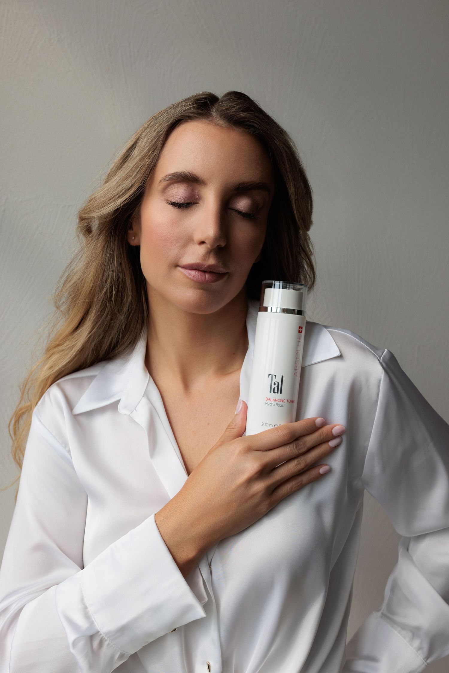 Blonde woman in white shirt holding Tal skincare bottle with eyes closed in serene pose.