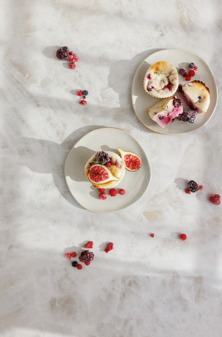 Two white plates with berry muffins and fig slices on marble surface with scattered fresh berries.