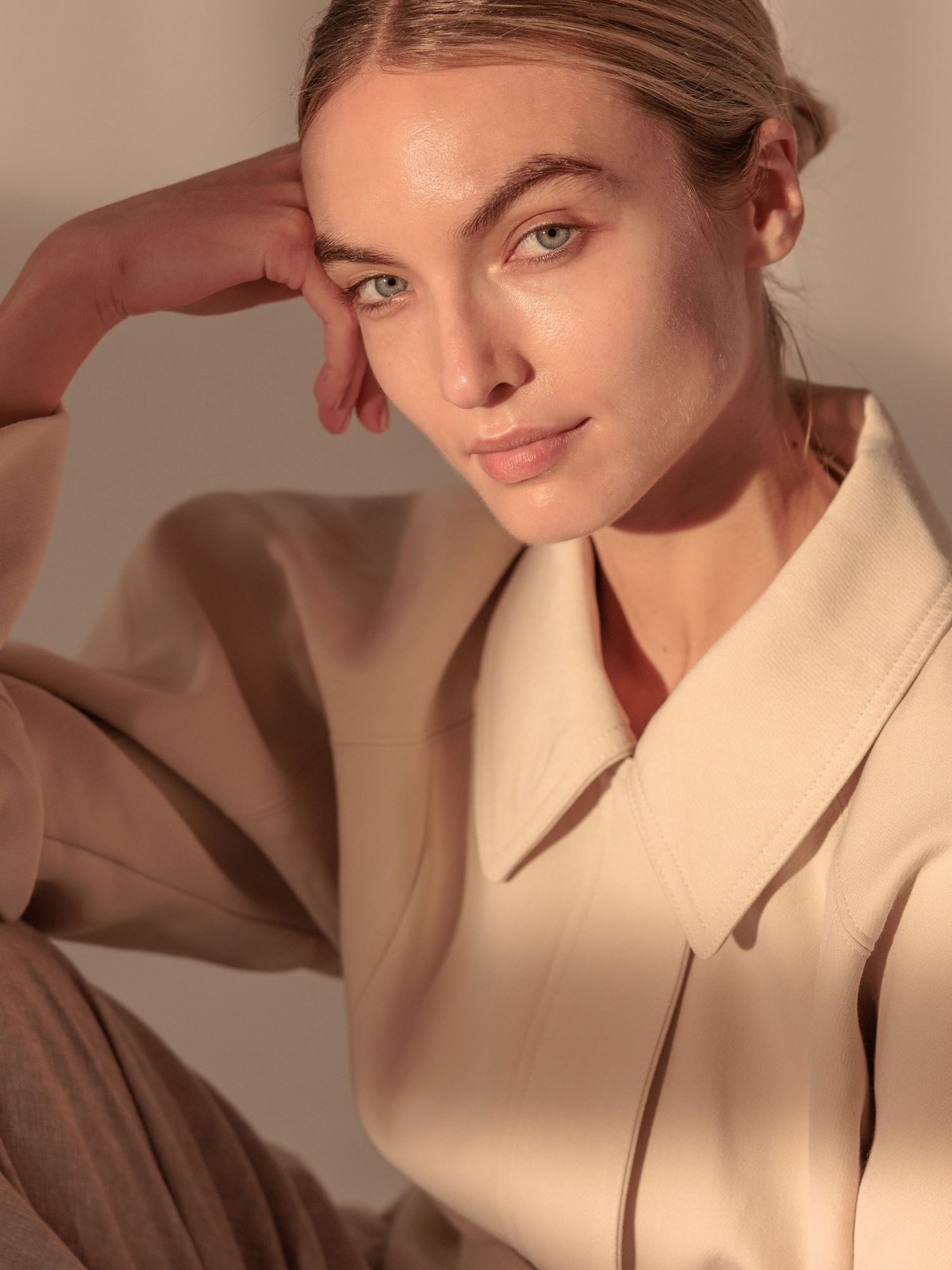 Woman in beige blazer poses with hand near face in warm natural light fashion portrait.