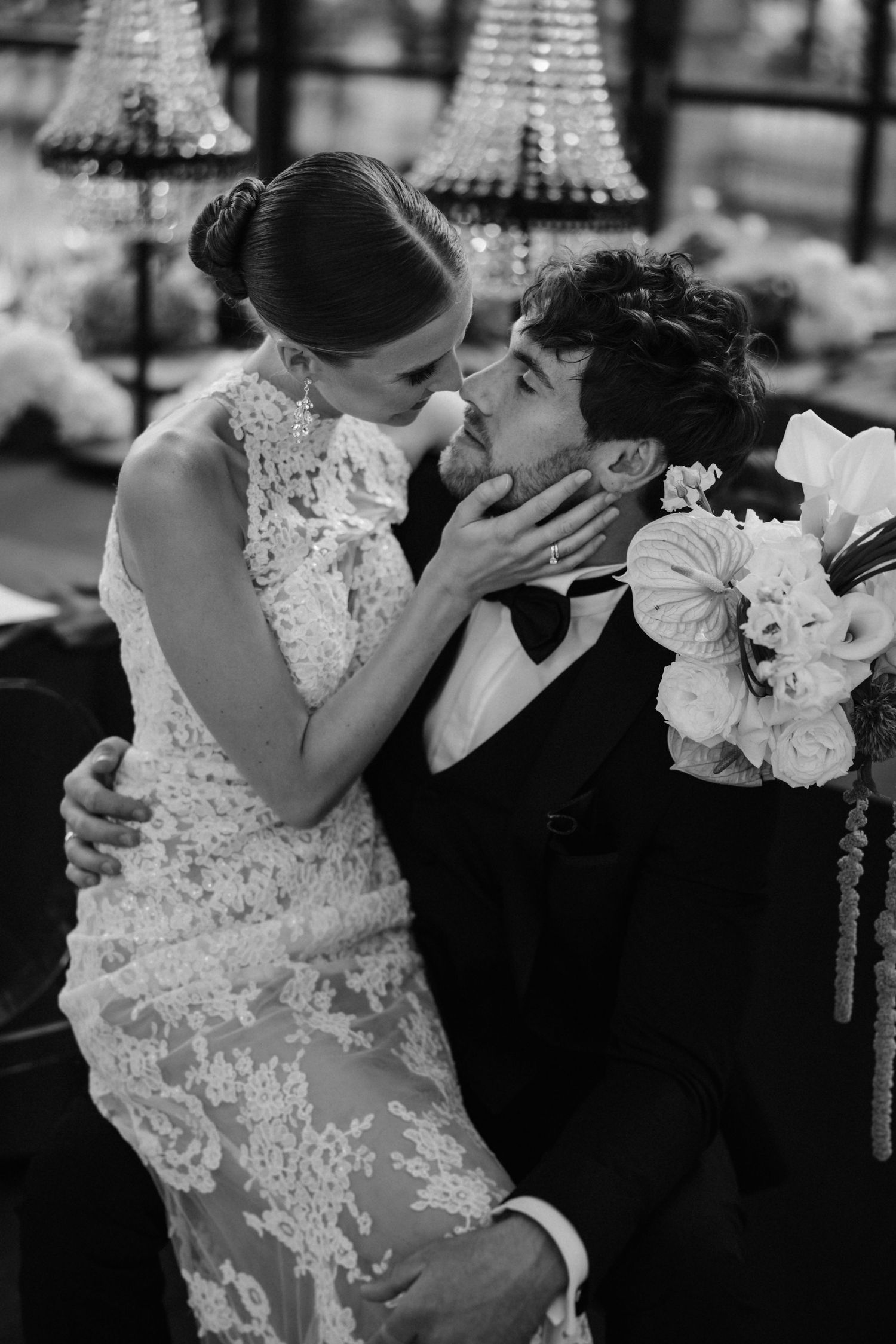 Black and white photo of bride in lace dress and groom in tuxedo sharing romantic moment.