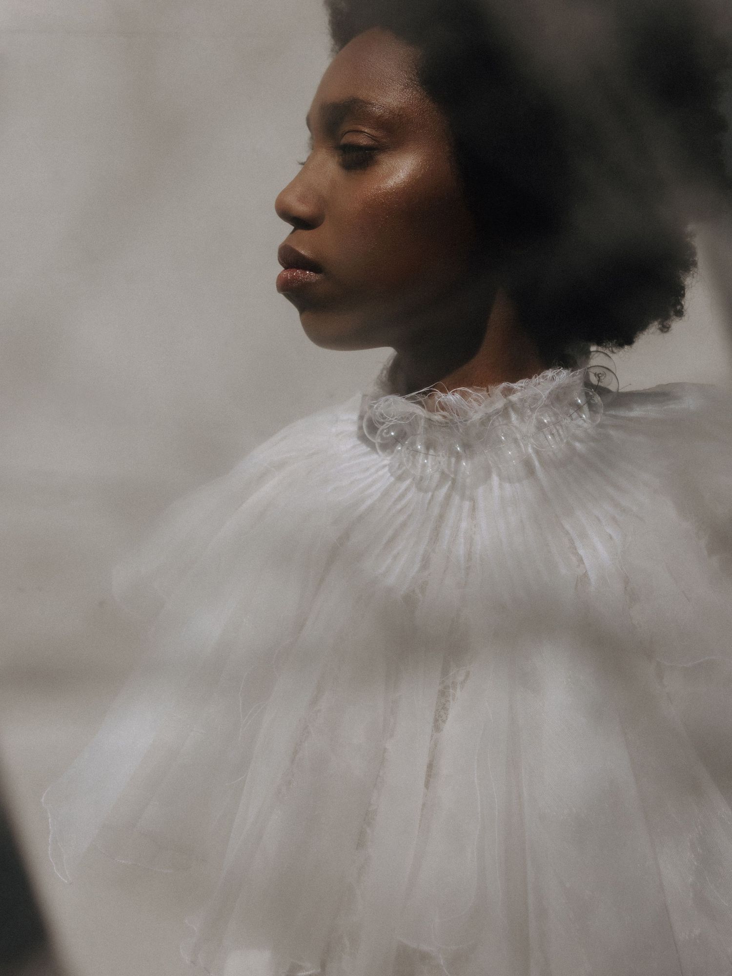 A Black woman in a white ruffled tulle cape gazes sideways in a dreamy, soft-focus editorial fashion portrait.