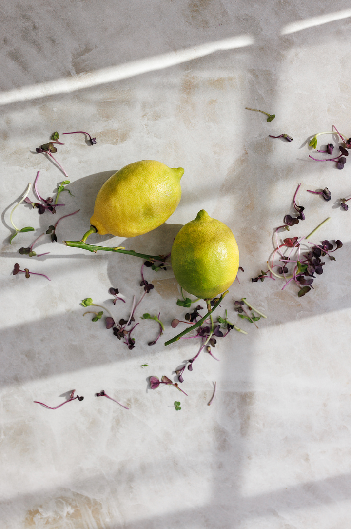 Two fresh yellow lemons surrounded by purple microgreens on marble surface in natural sunlight.