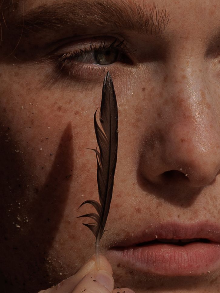 Close-up of freckled face with hazel eyes holding a dark feather against the cheek.