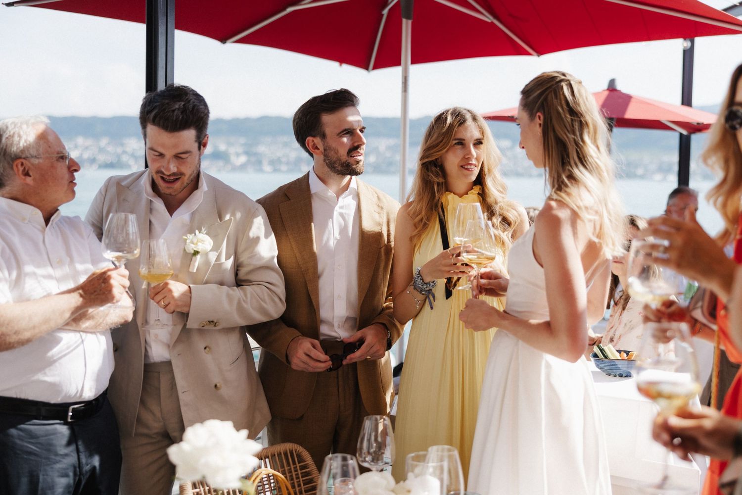 Elegantly dressed wedding guests socializing under red umbrellas at a waterfront reception.