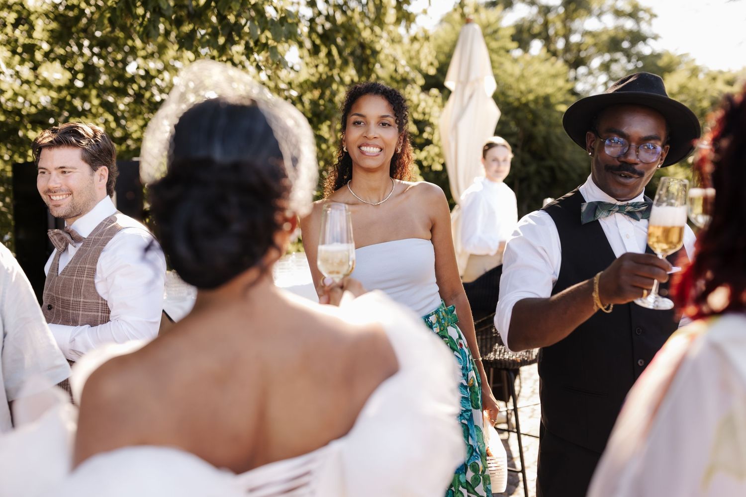 Diverse wedding guests socializing outdoors, smiling and toasting with champagne glasses in sunny garden setting.