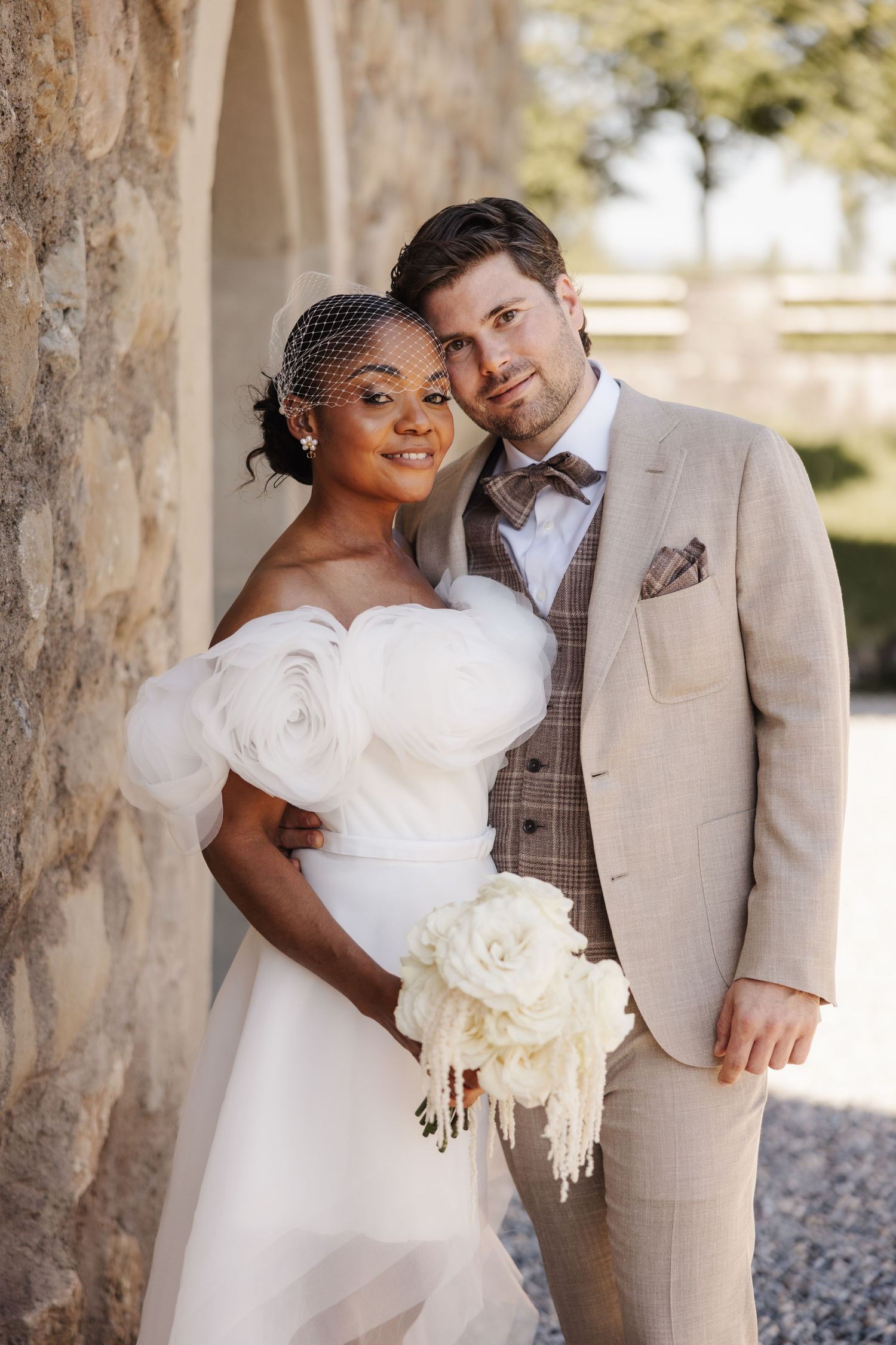 Elegant interracial bride and groom posing together beside a stone wall with white floral bouquet.