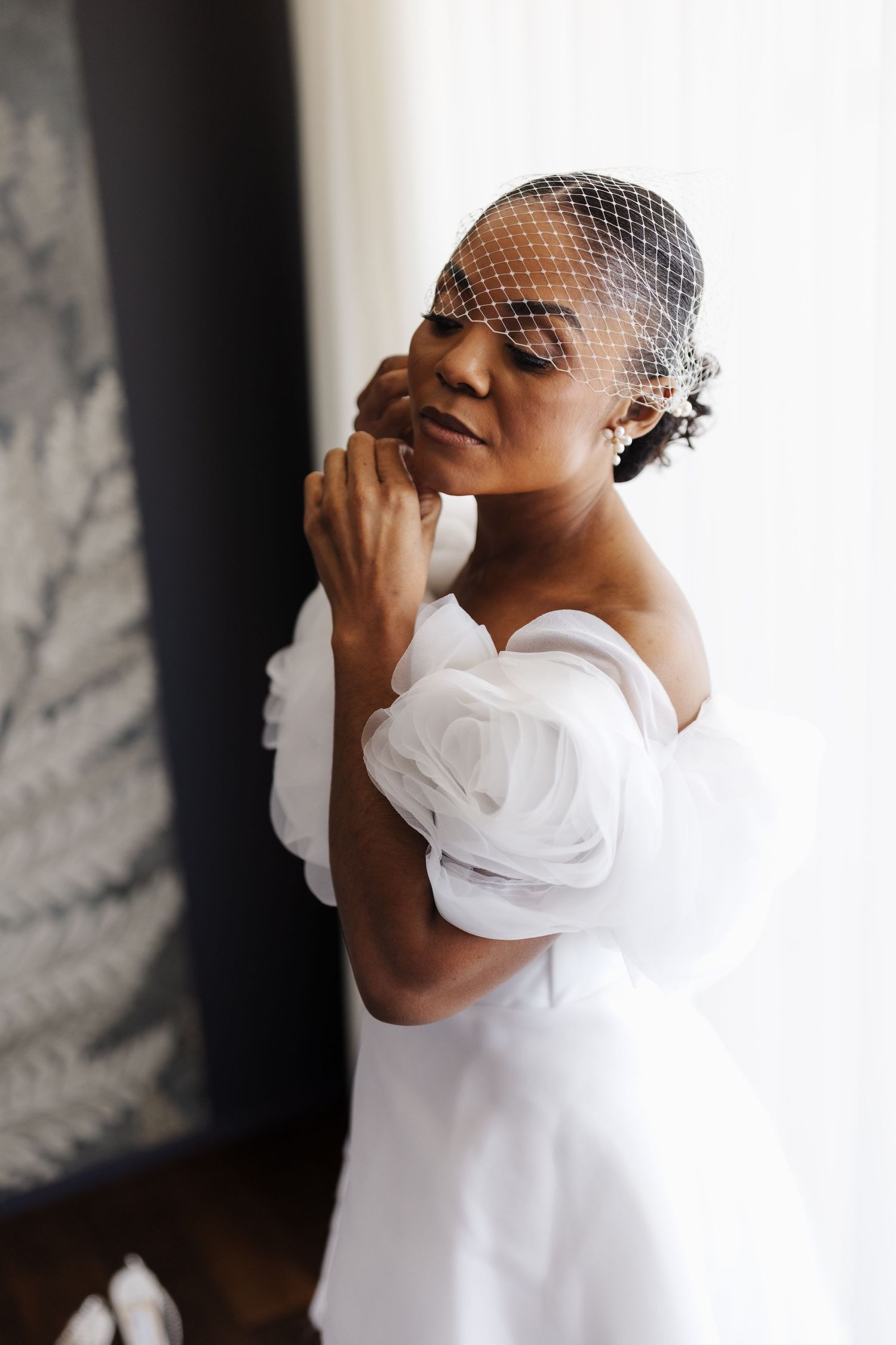 Elegant Black bride in white off-shoulder tulle gown and birdcage veil adjusts her earring by window.