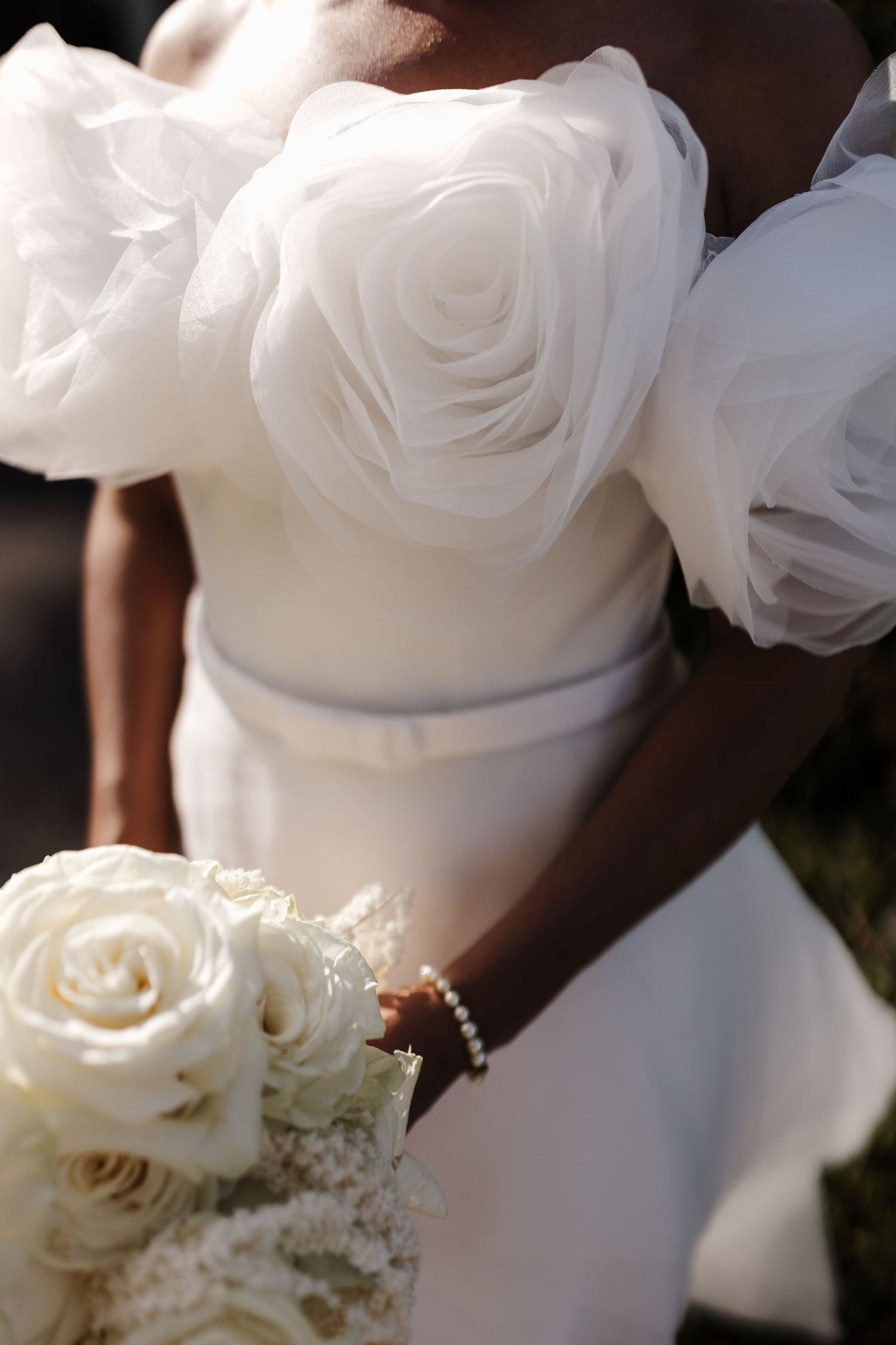 Bride in elegant white off-shoulder wedding gown with rose details holding a white rose bouquet with pearl bracelet.