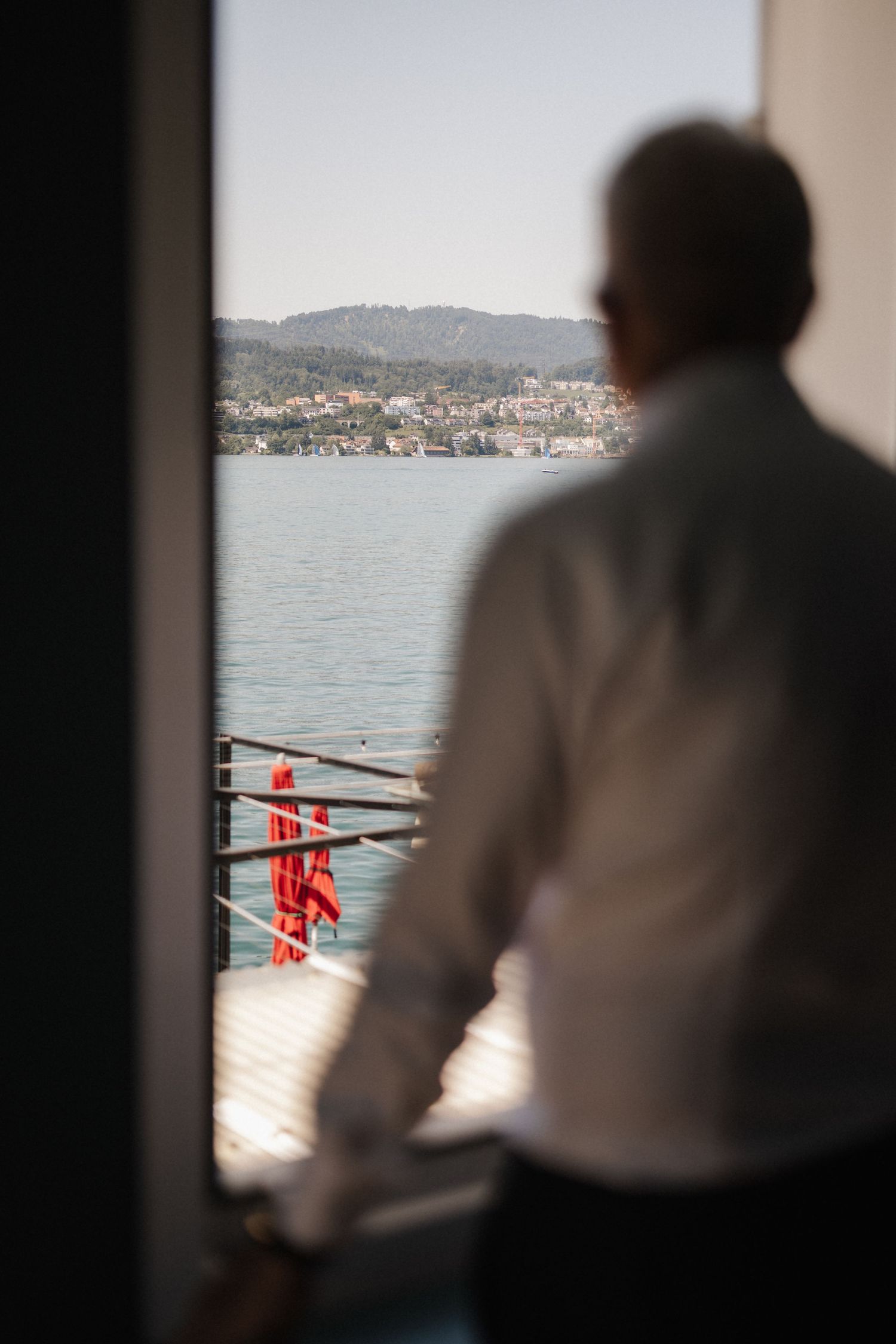 Man in white shirt standing on a balcony overlooking a scenic waterfront town on a sunny day.