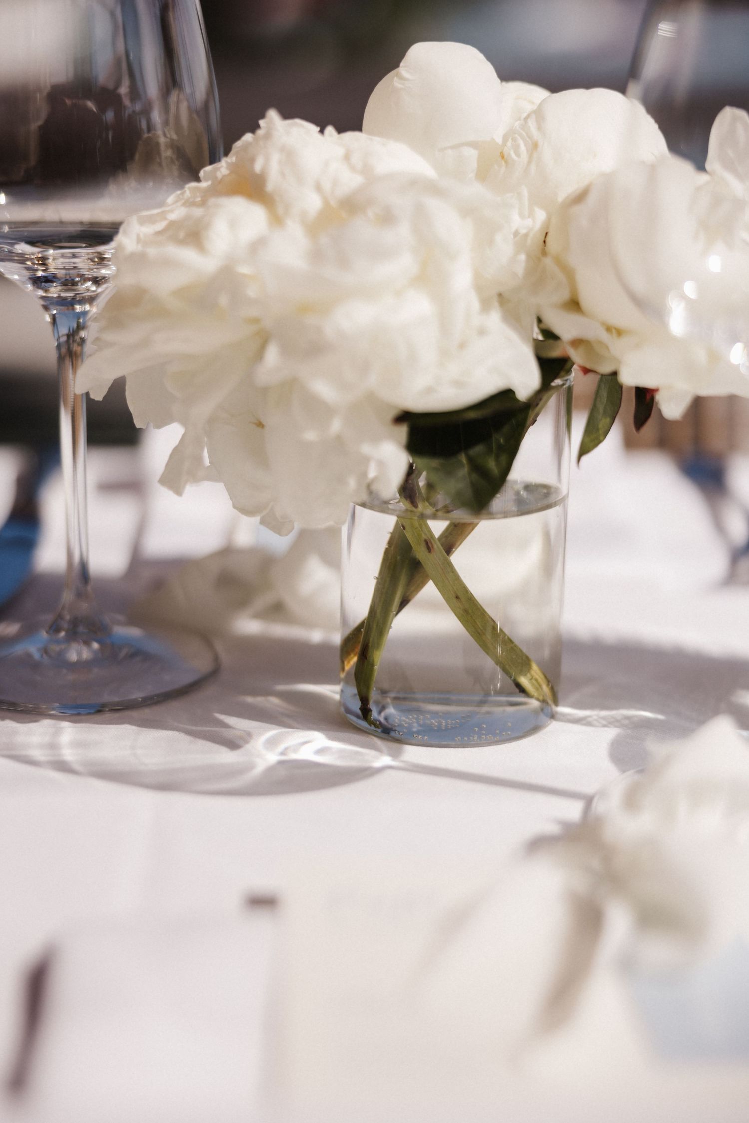 White peony flowers in a clear glass vase on an elegant white wedding reception table with wine glasses.