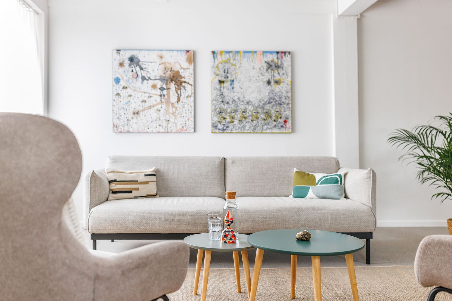 Minimalist living room featuring gray sectional sofa, teal coffee tables, and abstract paintings.