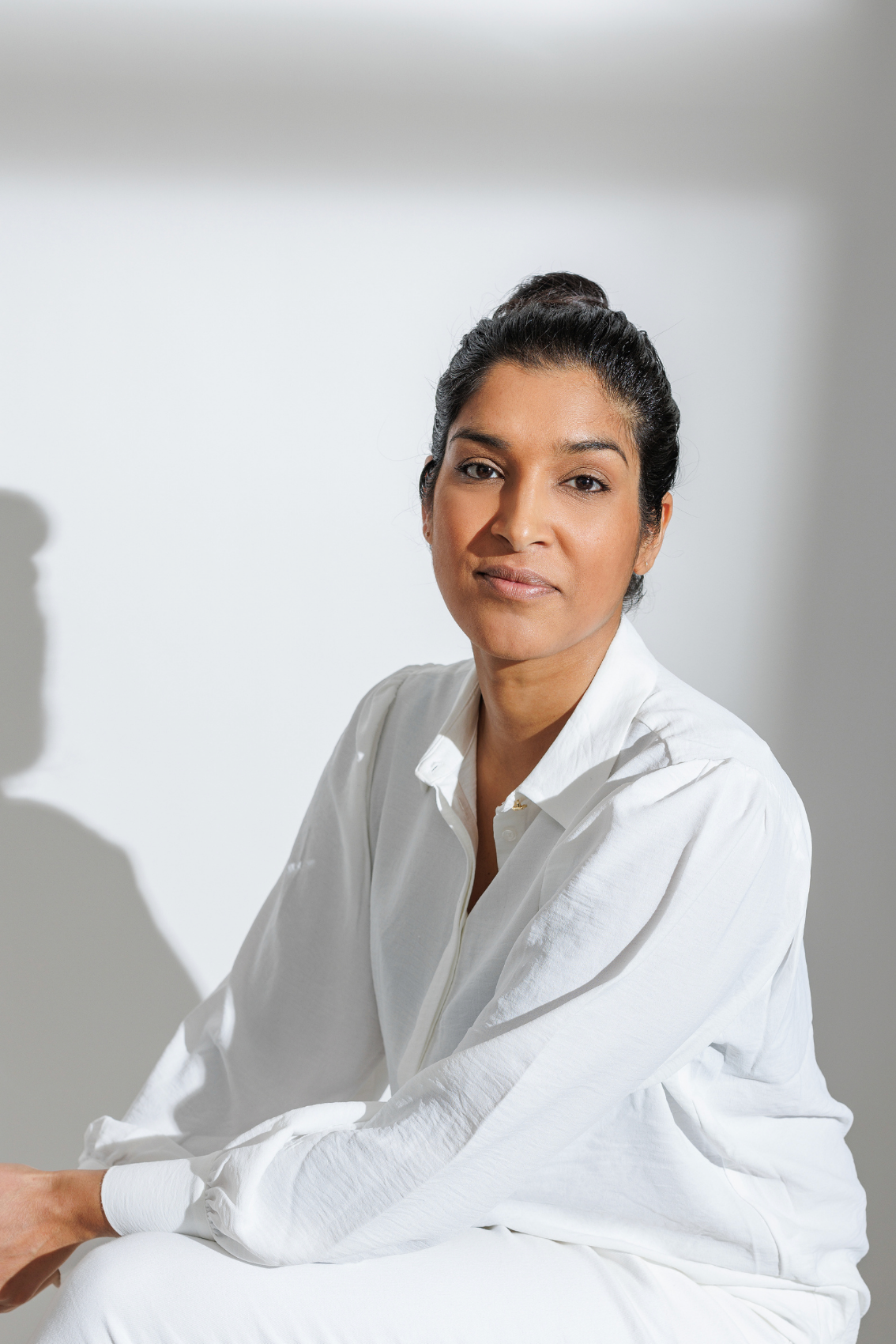 A woman in a white linen shirt sits calmly against a white wall in natural sunlight.