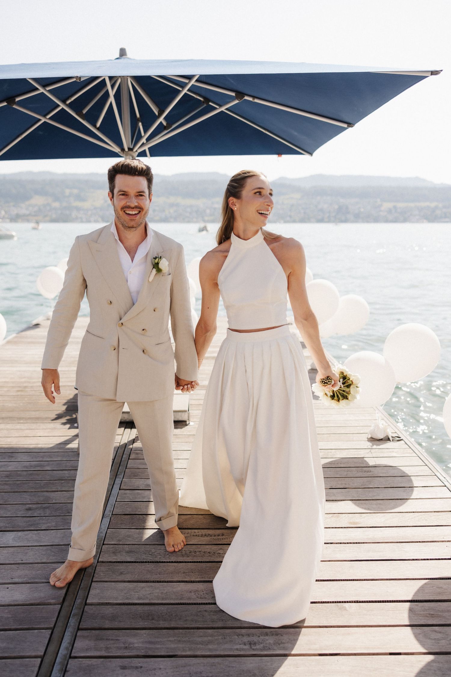 Happy bride and groom holding hands walking barefoot on a wooden dock by a scenic lake.