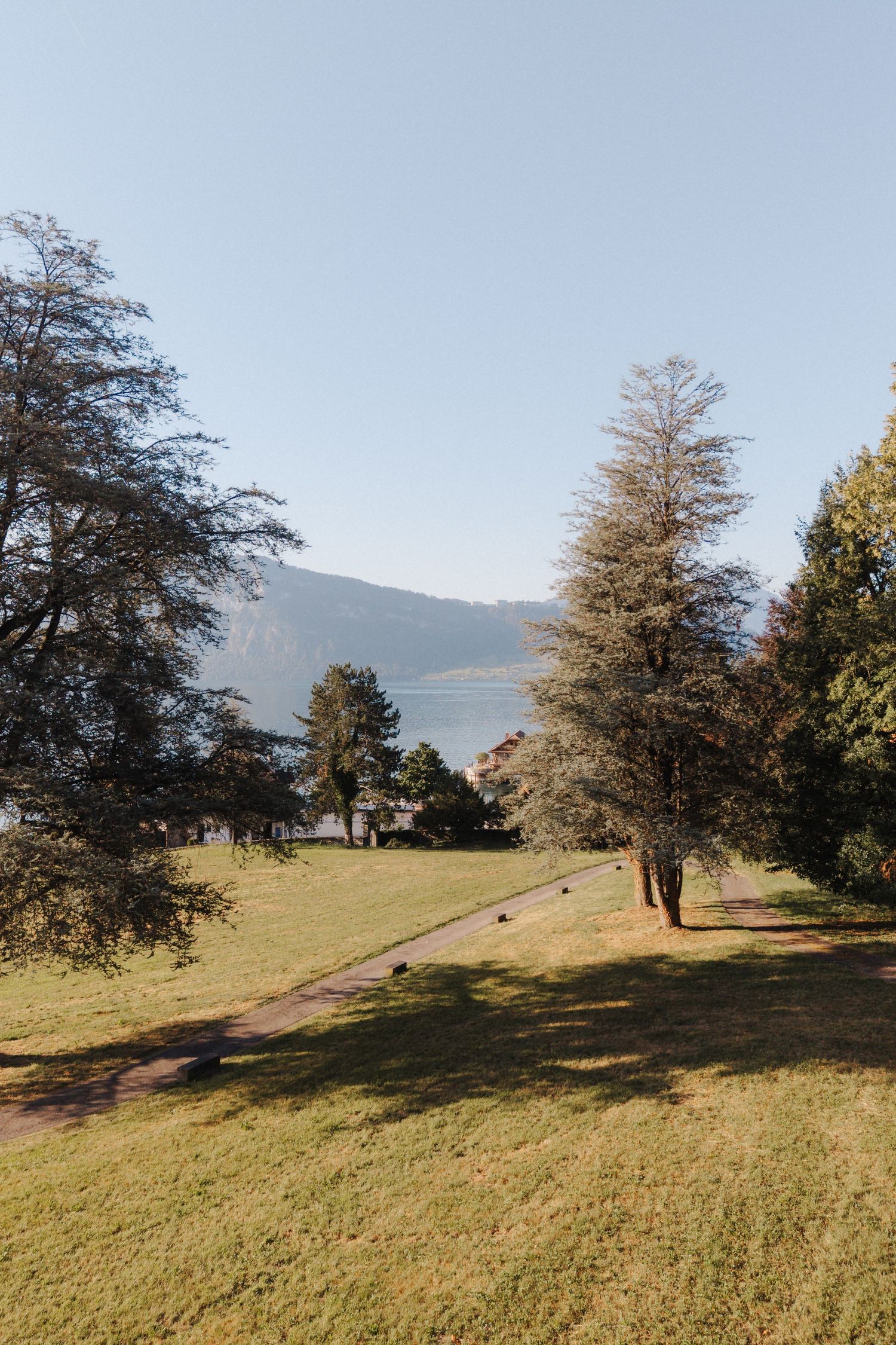 Tree-lined pathway through green grounds with a scenic mountain lake visible in the background.