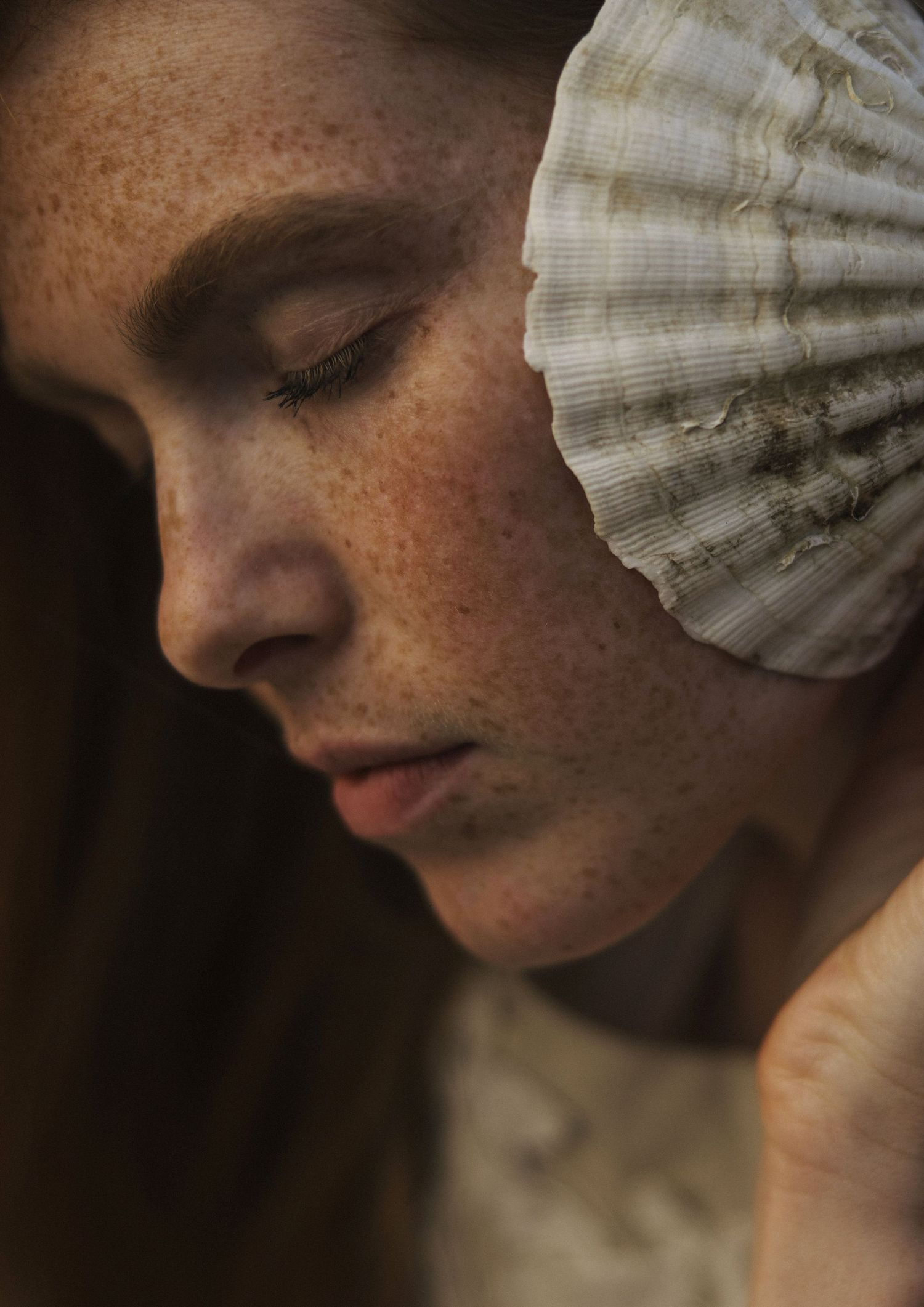 Close-up portrait of a freckled woman holding a seashell against her cheek with eyes closed.