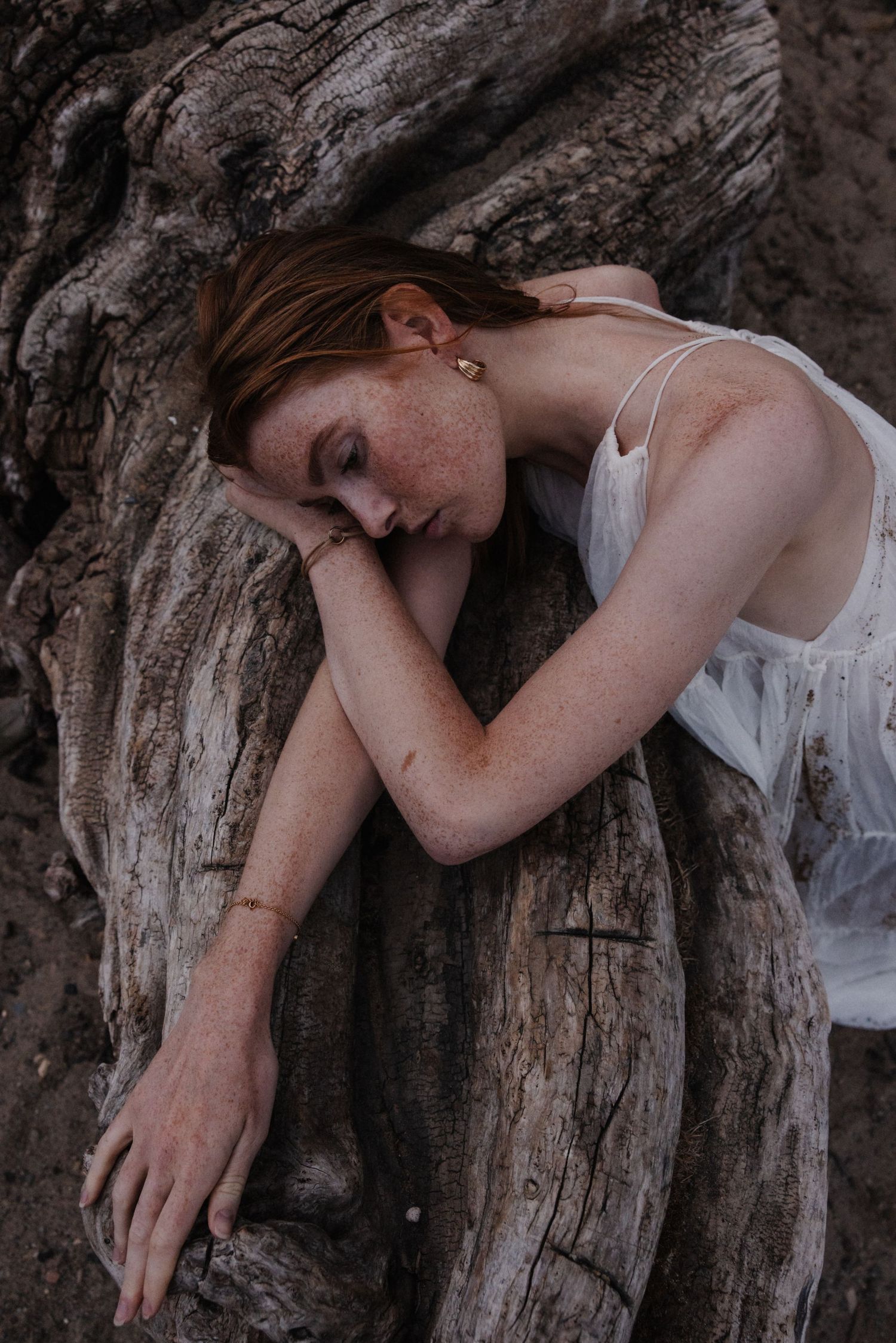 Red-haired freckled woman in white dress resting against weathered driftwood roots.