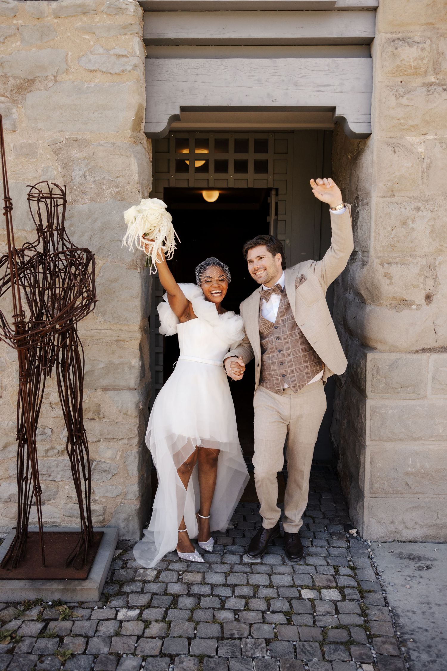 Joyful interracial newlyweds celebrating as they exit stone building doorway after wedding ceremony.