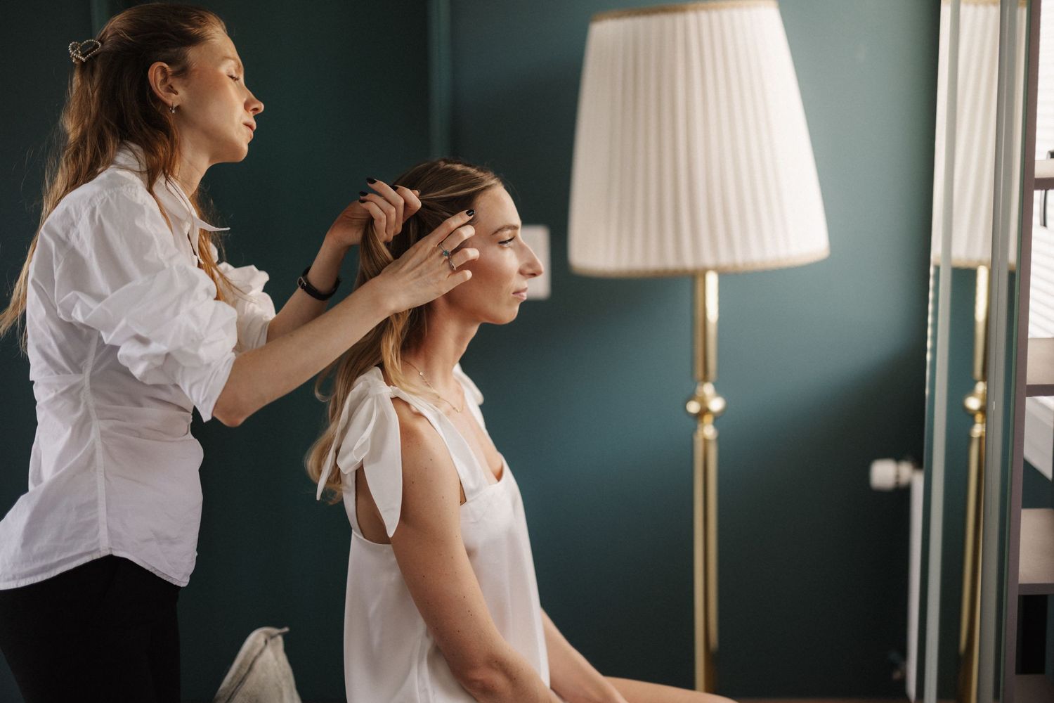 Female hairstylist styling seated bride's hair near lamp in teal room.