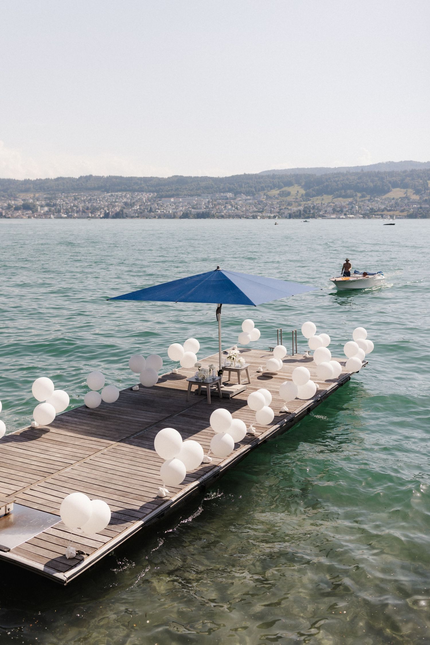 Wooden dock decorated with white balloons and blue umbrella on a calm lake with a boat passing by.