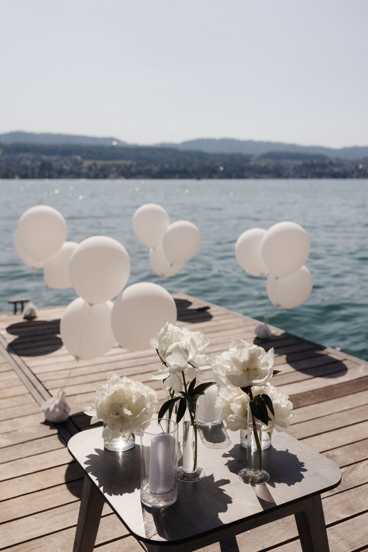 White balloons and peony flowers in vases on a wooden pier overlooking a sparkling blue lake.