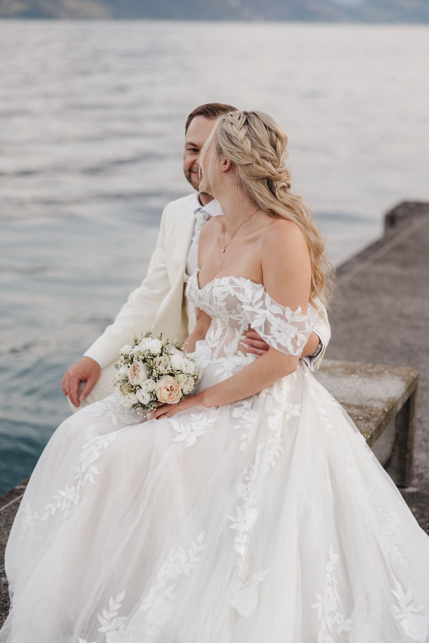 Laughing bride in lace off-shoulder gown holding bouquet seated with groom on a dock overlooking a lake.