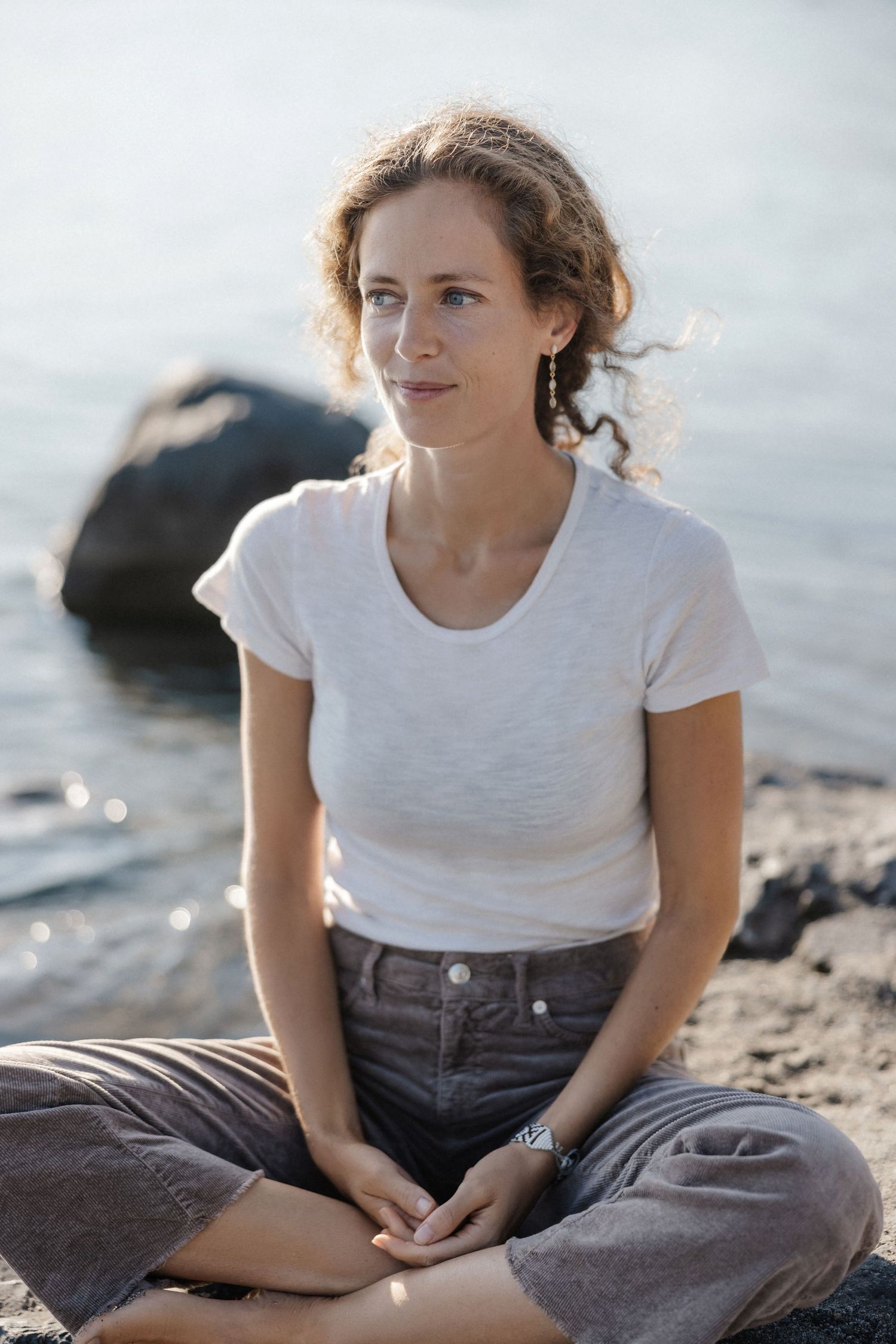 Woman in white t-shirt and gray pants sitting cross-legged on rocks by a shimmering body of water.